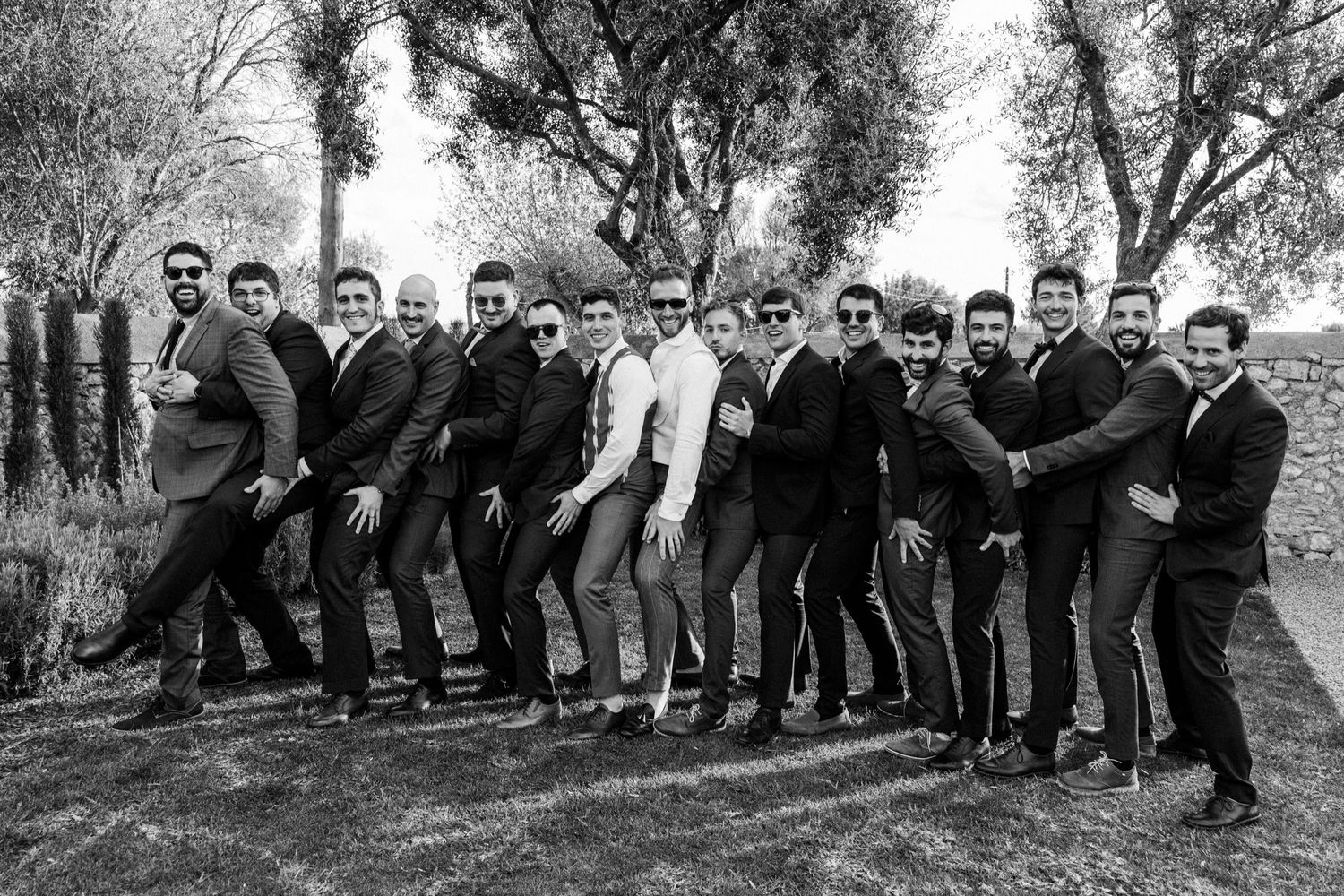 Wedding party members pose in a casual line formation on a grassy slope outdoors.