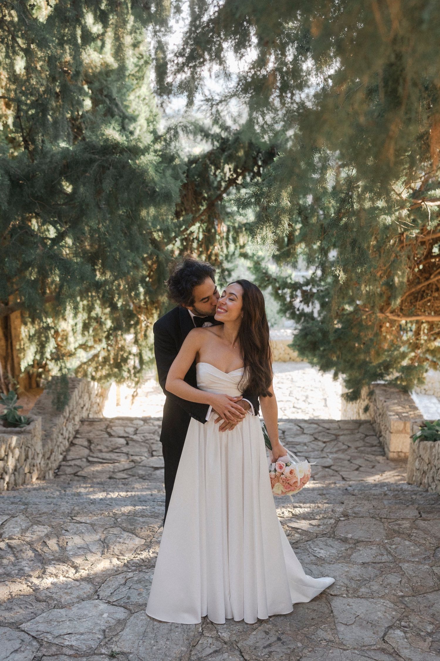 A couple embraces on a stone patio surrounded by evergreen trees during a romantic outdoor wedding photoshoot.