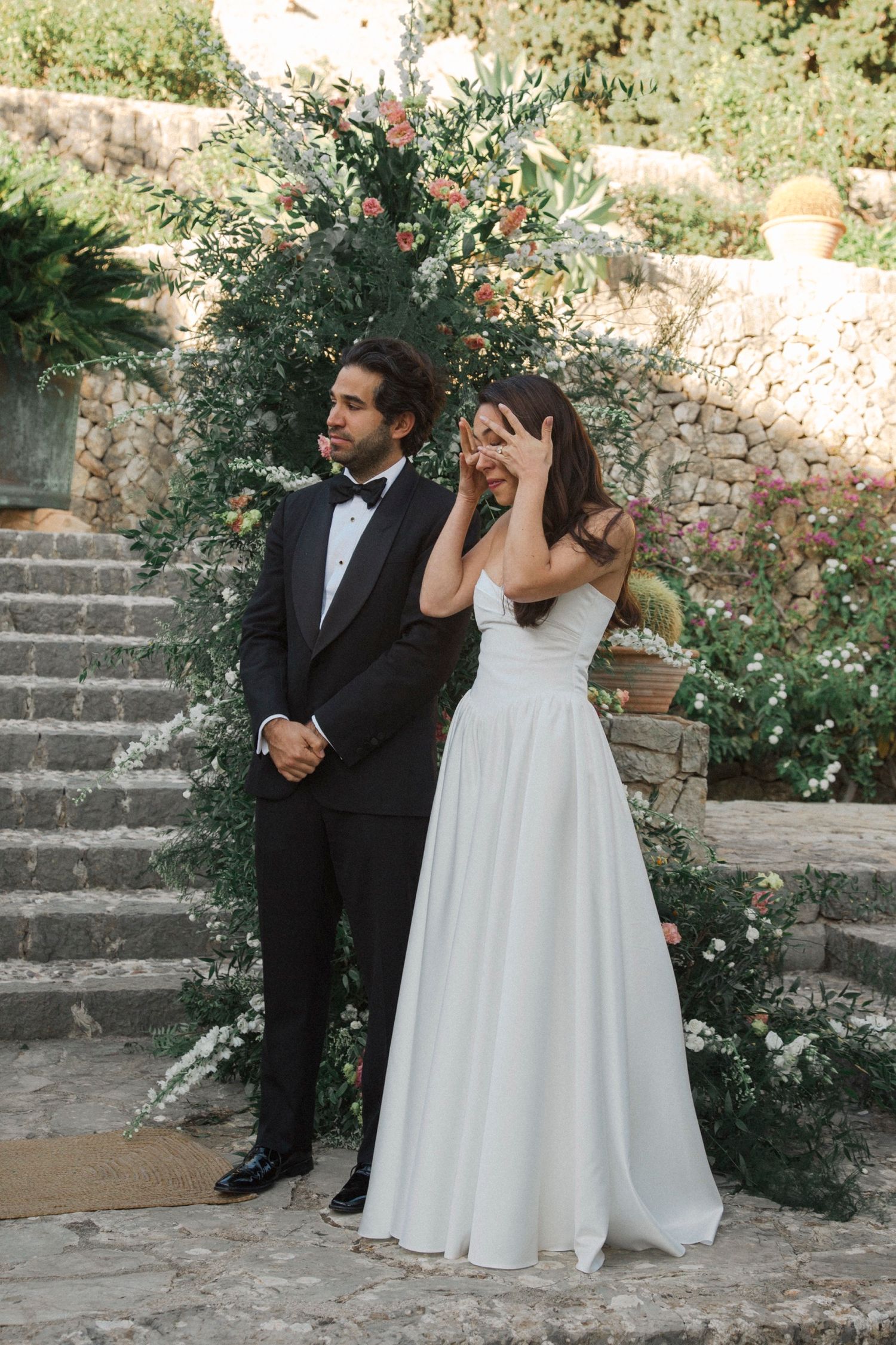 A newlywed couple shares candid moments on stone steps surrounded by blooming flowers in a garden setting.