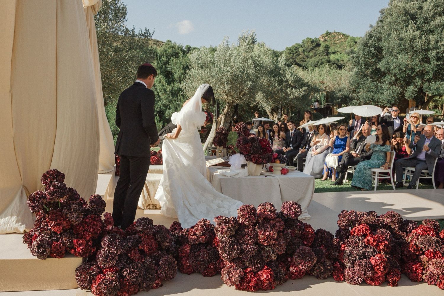 Wedding guests gather on an outdoor patio surrounded by lush greenery and red floral arrangements.