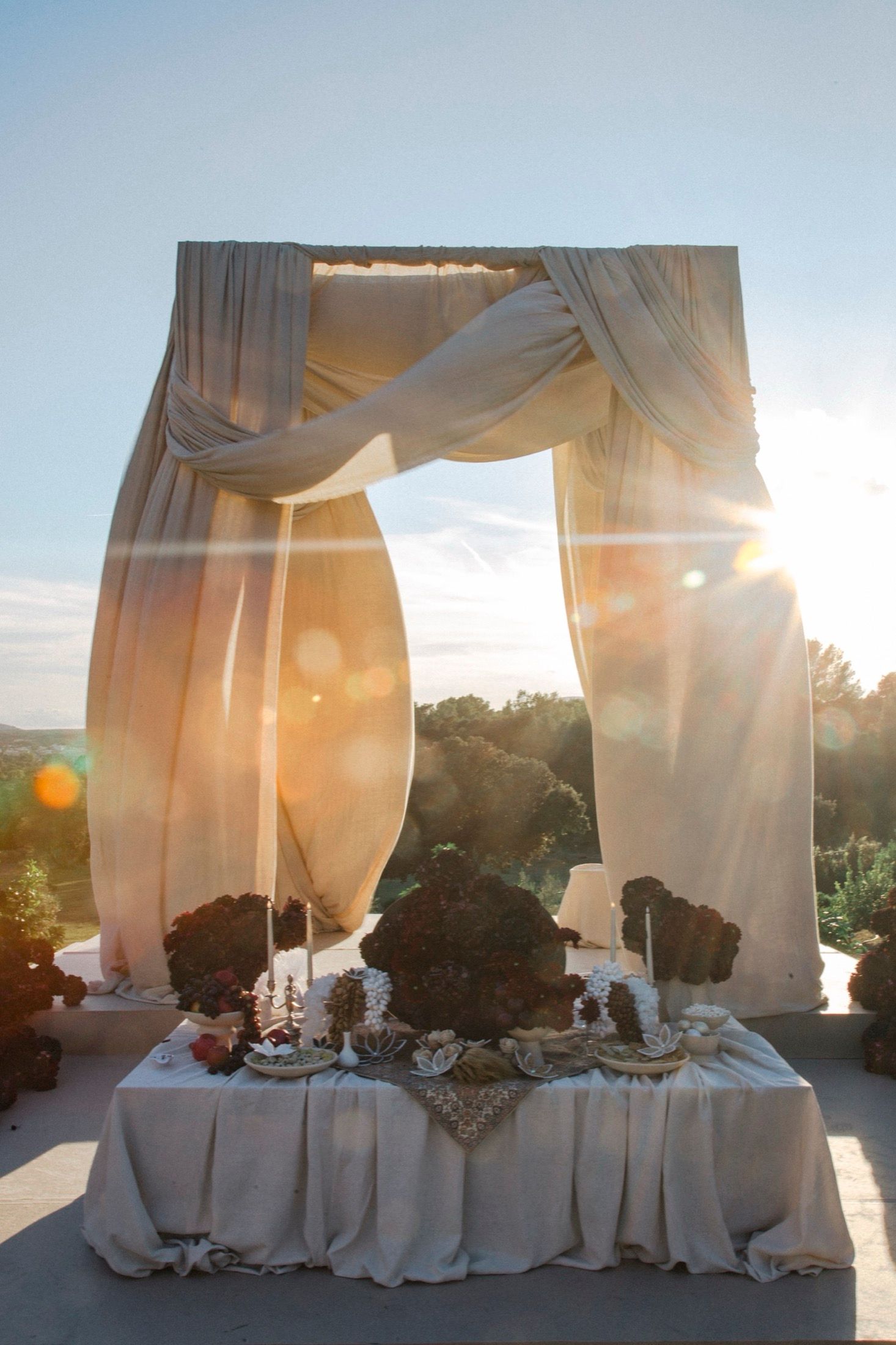 Elegant outdoor dining table setup with flowing white curtains and golden sunlight streaming through at sunset.