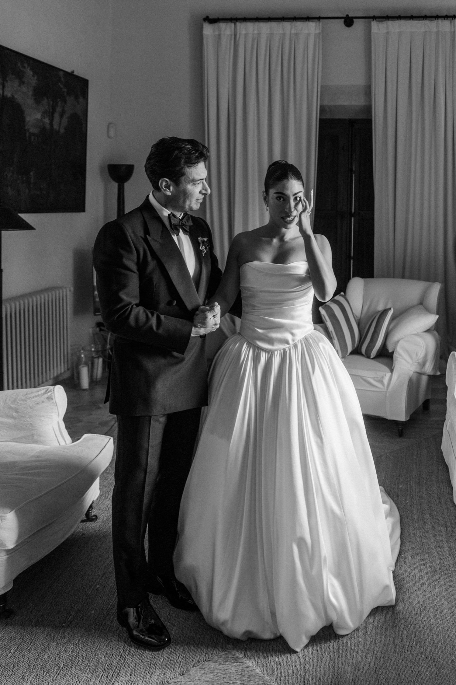 A black and white wedding photo captures a tender moment between a couple in formal attire in a living room setting.