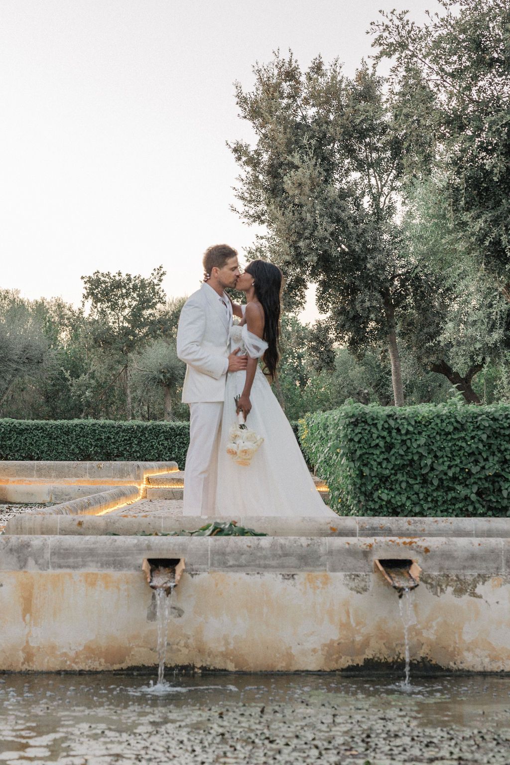 Romantic wedding couple standing by a fountain at sunset in an elegant garden setting.