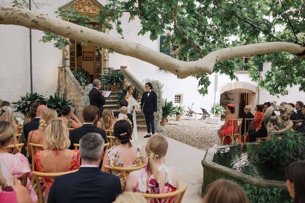 An outdoor wedding ceremony with guests seated beneath a large tree in a courtyard.