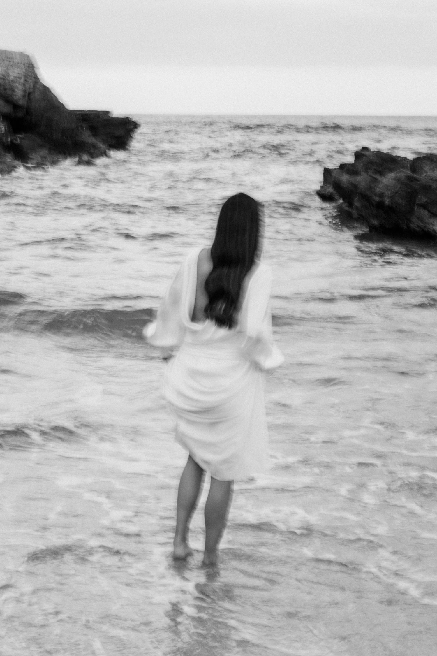 Black and white image of person standing in ocean waves wearing flowing white dress.