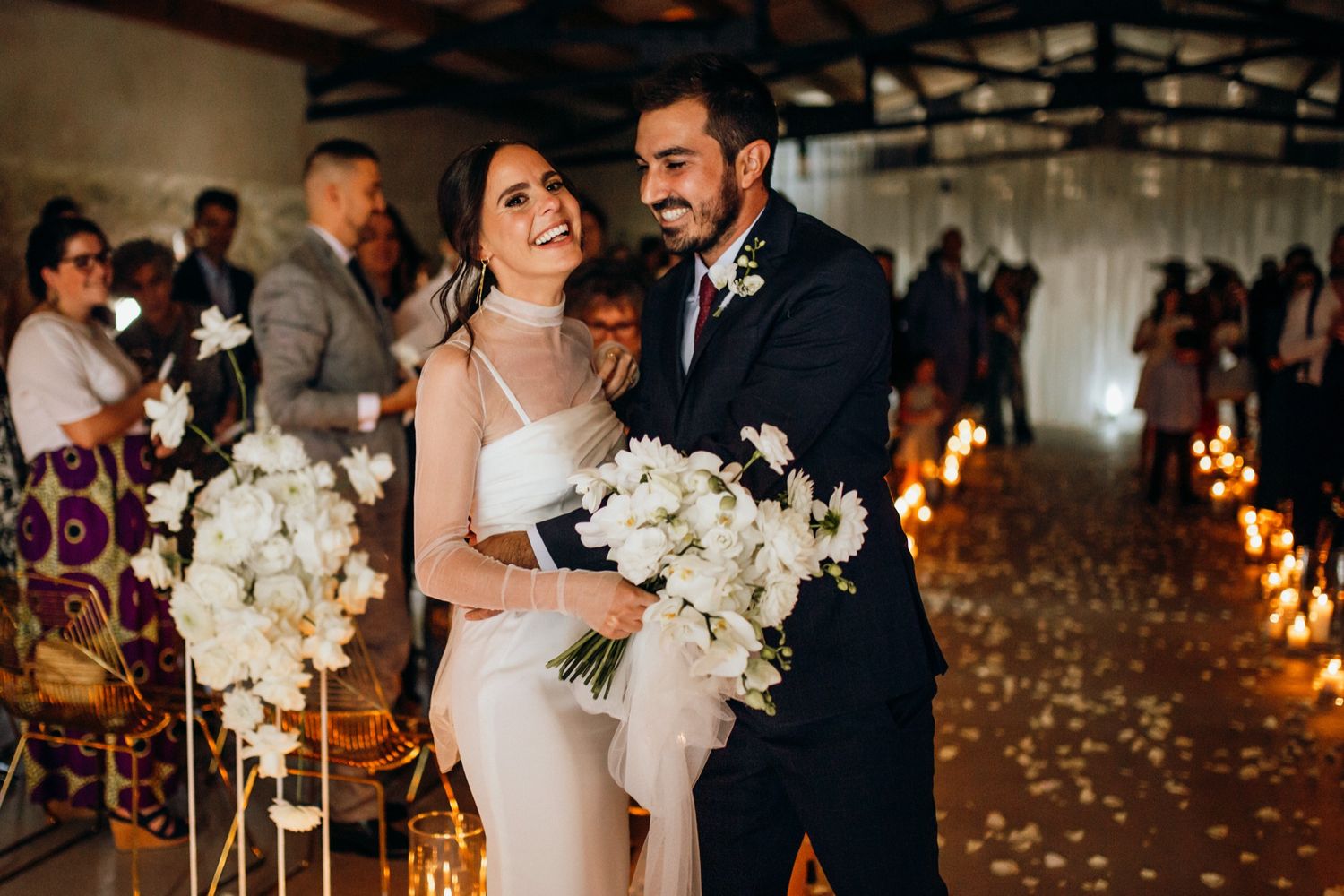 A couple shares a joyful moment at their candlelit wedding ceremony with white orchid bouquets and rose petals on the floor.
