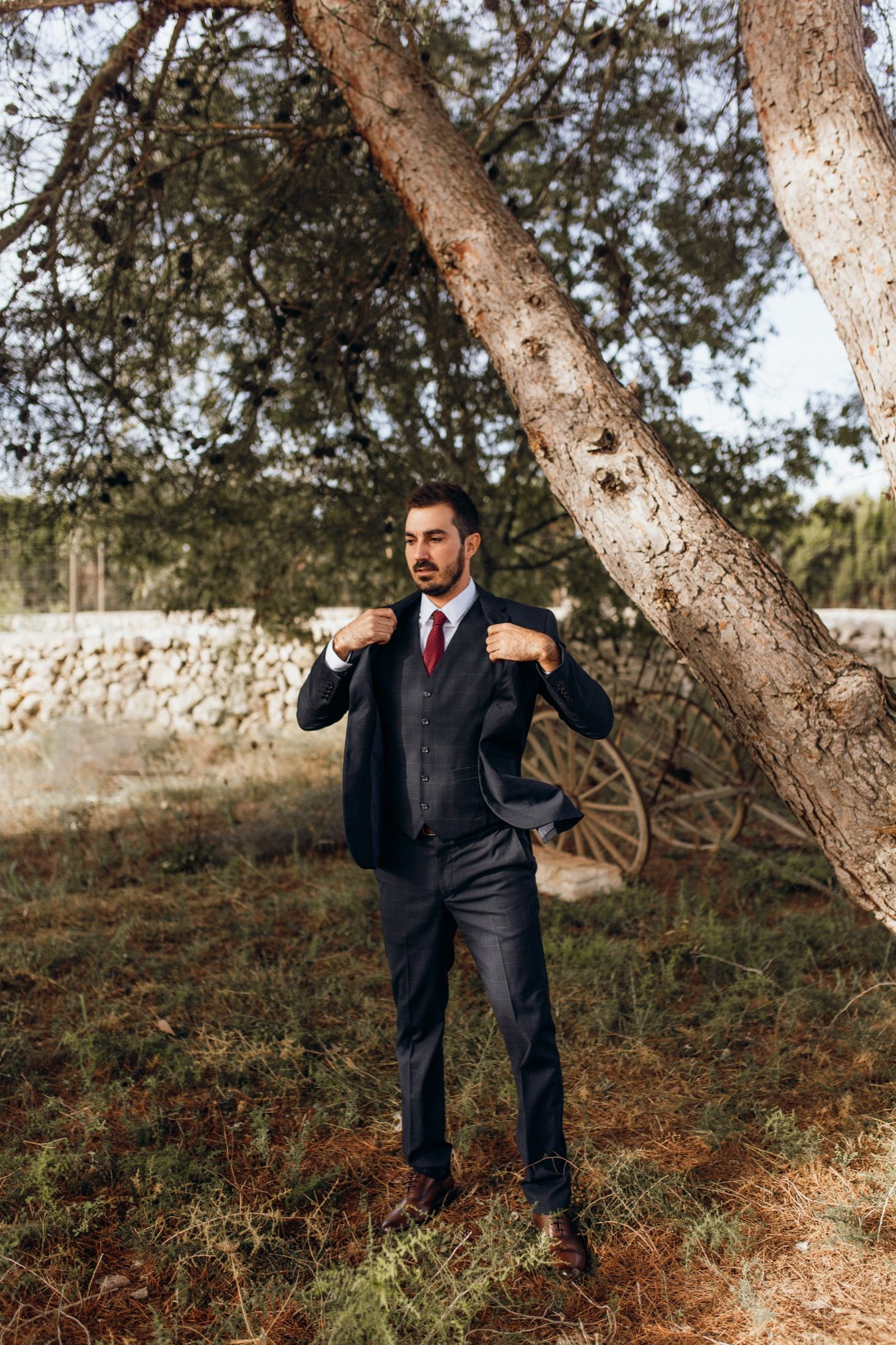 A person in a dark suit poses playfully near a tree trunk in an outdoor autumn setting.