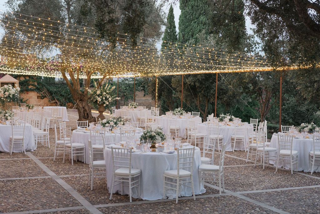 Outdoor wedding reception setup with white chairs and tables under string lights at dusk.