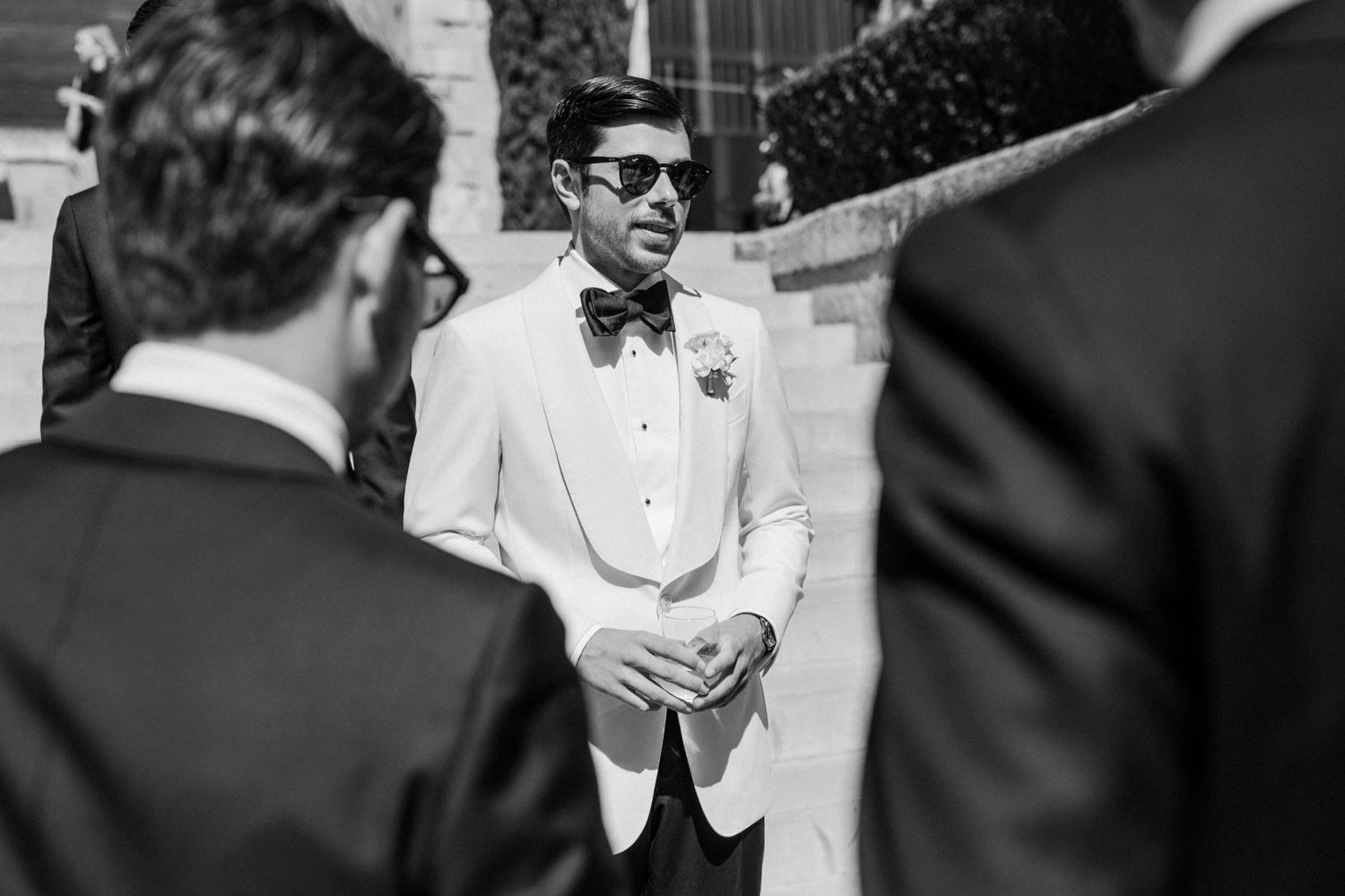 Black and white photo of a groom in white tuxedo and bow tie wearing sunglasses at an outdoor wedding ceremony.