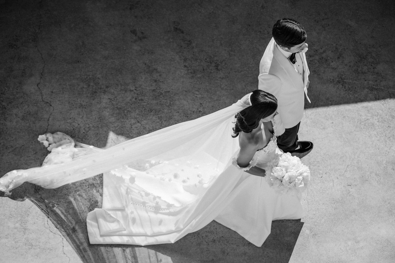 Aerial black and white photo of a bride and groom sharing a kiss, with elegant wedding dress train spread out.