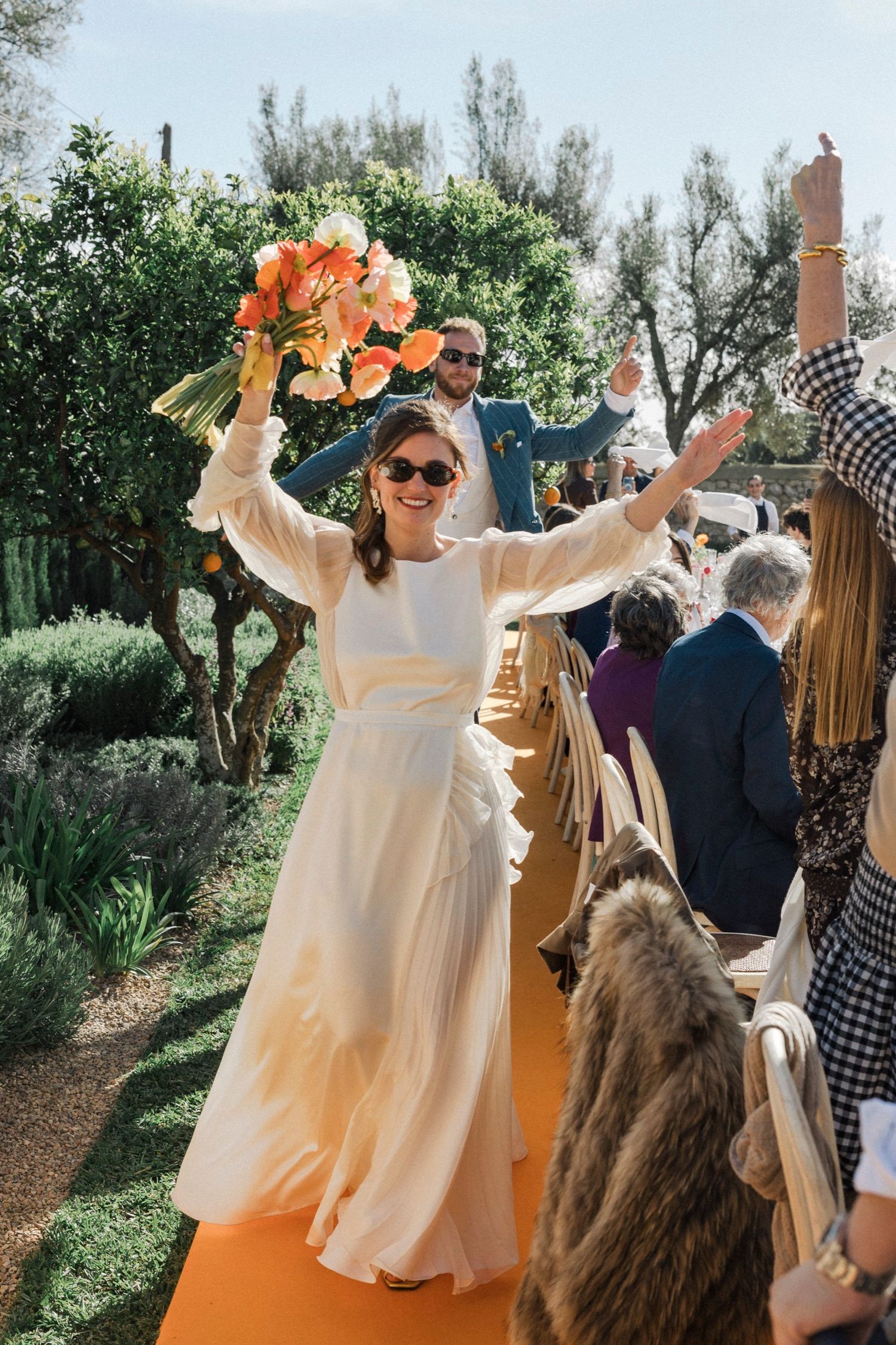 A person in a flowing cream-colored dress dances joyfully outdoors among trees and wedding guests.