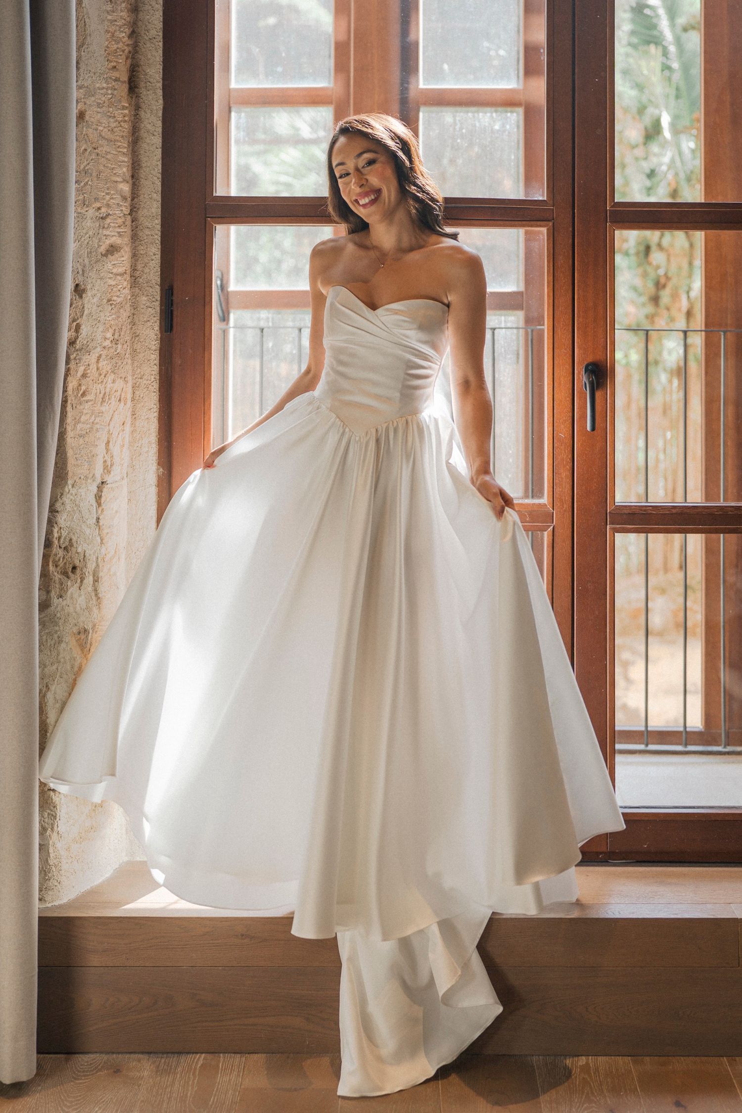 A stunning white wedding gown with a strapless sweetheart neckline and full ballgown skirt is showcased by a window.
