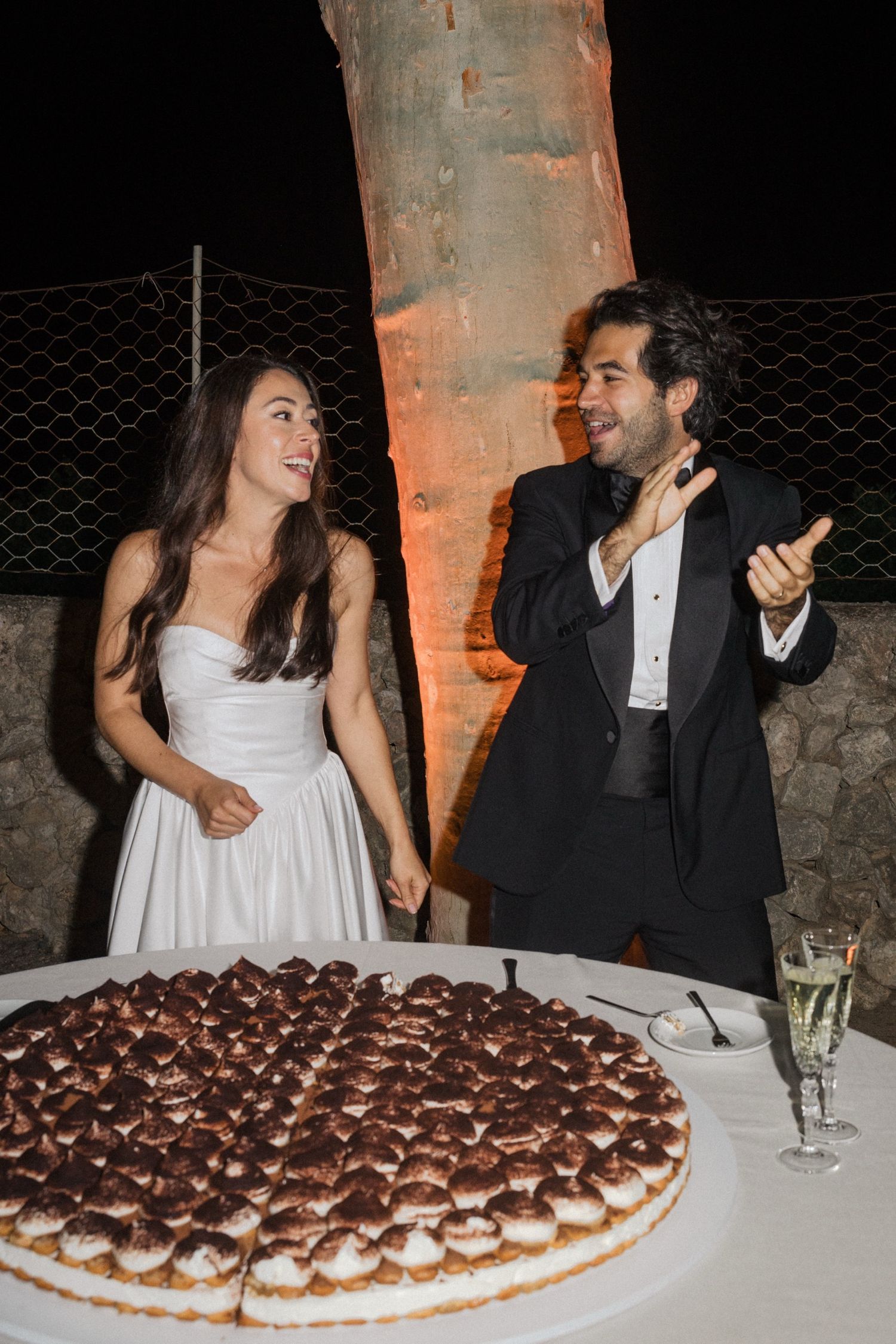 A couple laughs together at their wedding reception next to a large tiramisu dessert and champagne glass.