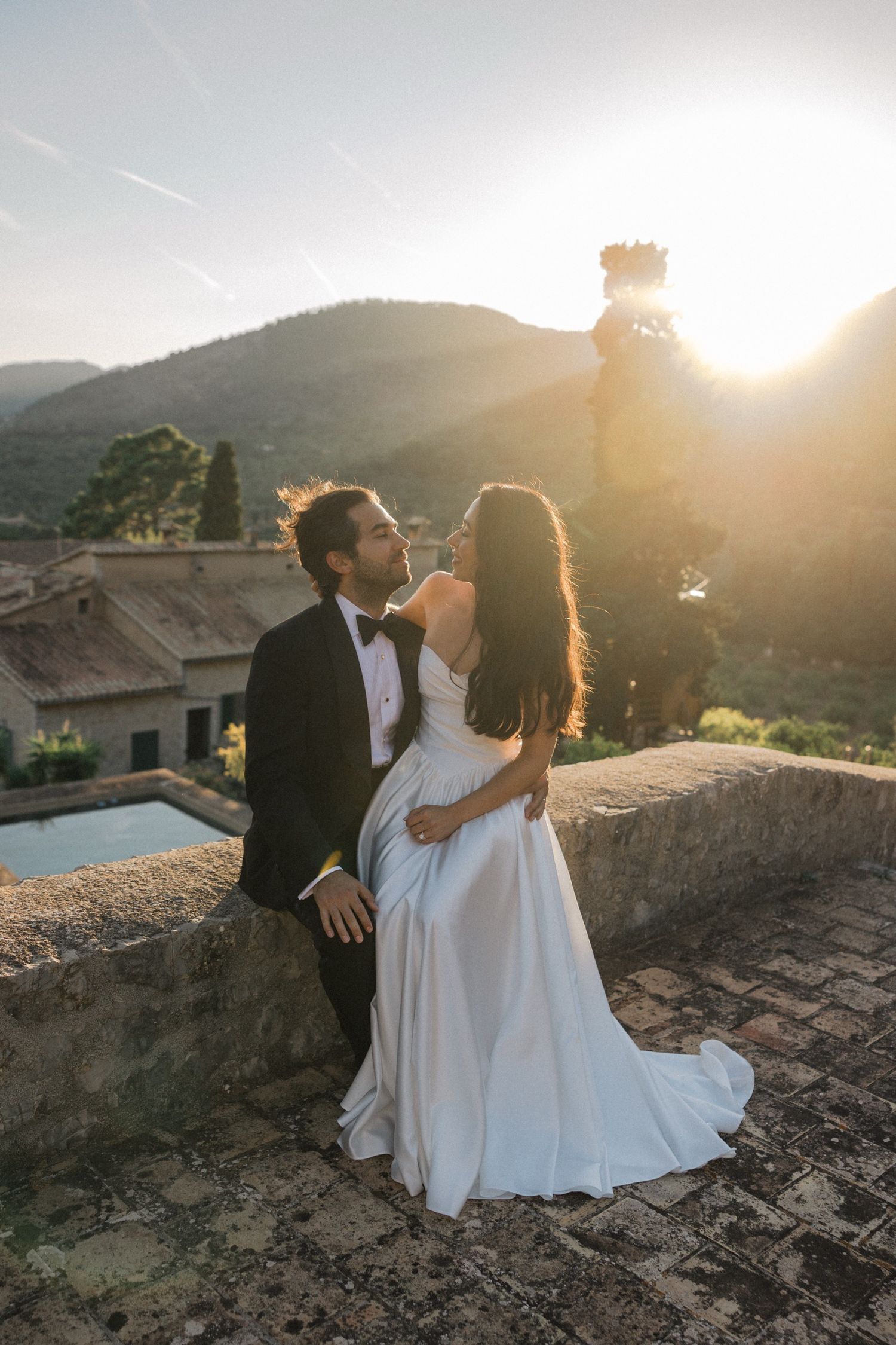 A couple in wedding attire embrace at sunset on a stone terrace overlooking mountains and a pool below.