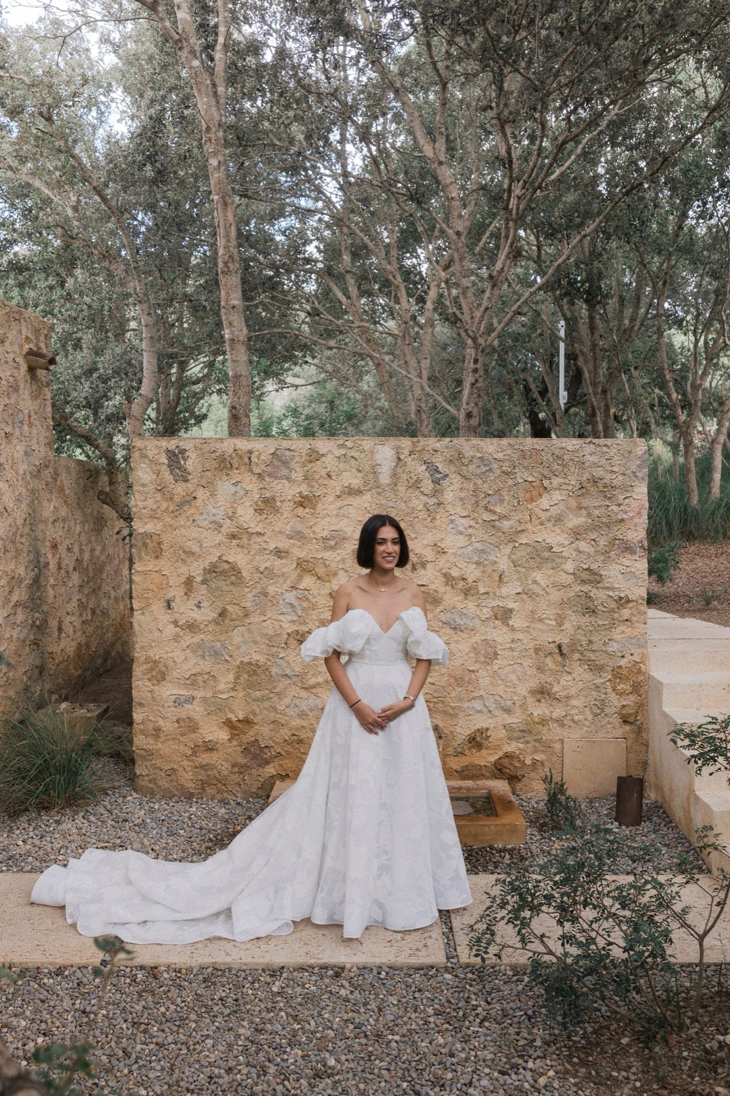 A stunning white off-shoulder wedding gown with a long train is showcased against a rustic stone wall in a garden setting.