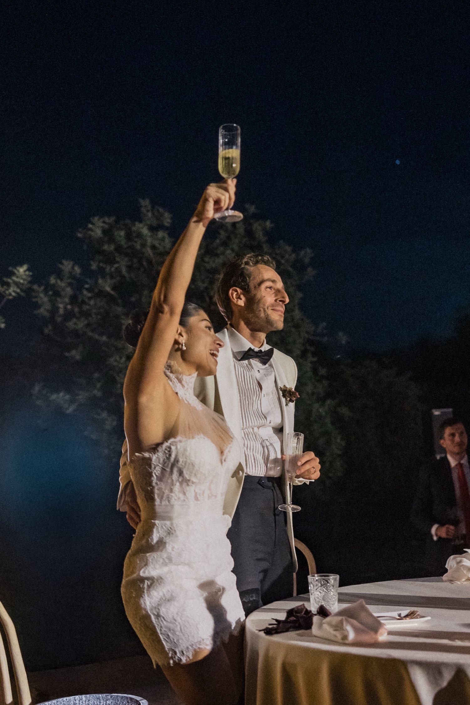 A bride in a white lace dress raises her arm in celebration during an evening outdoor wedding reception.