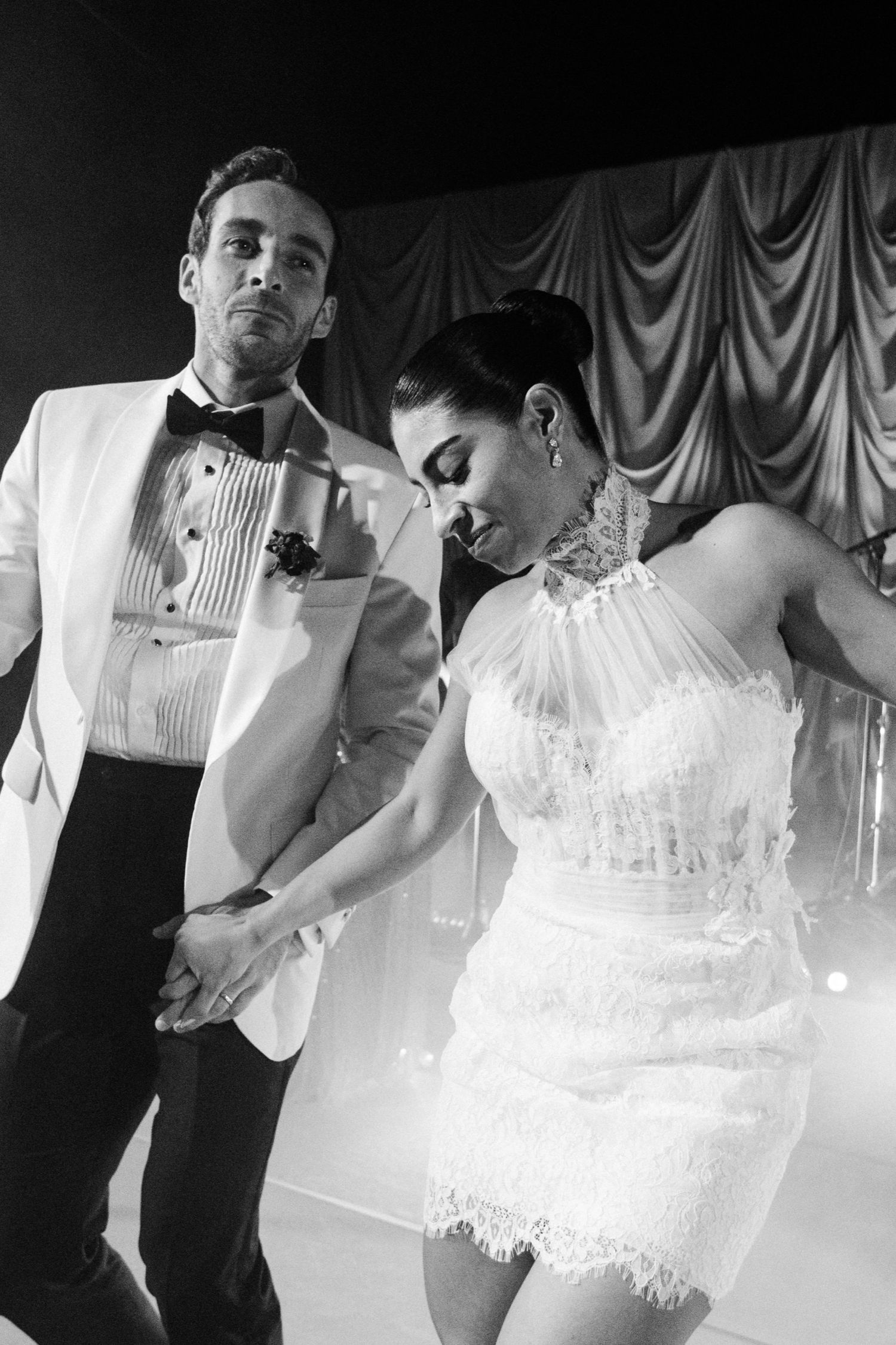Newlyweds share an intimate dance together, with the bride wearing a detailed white lace gown.