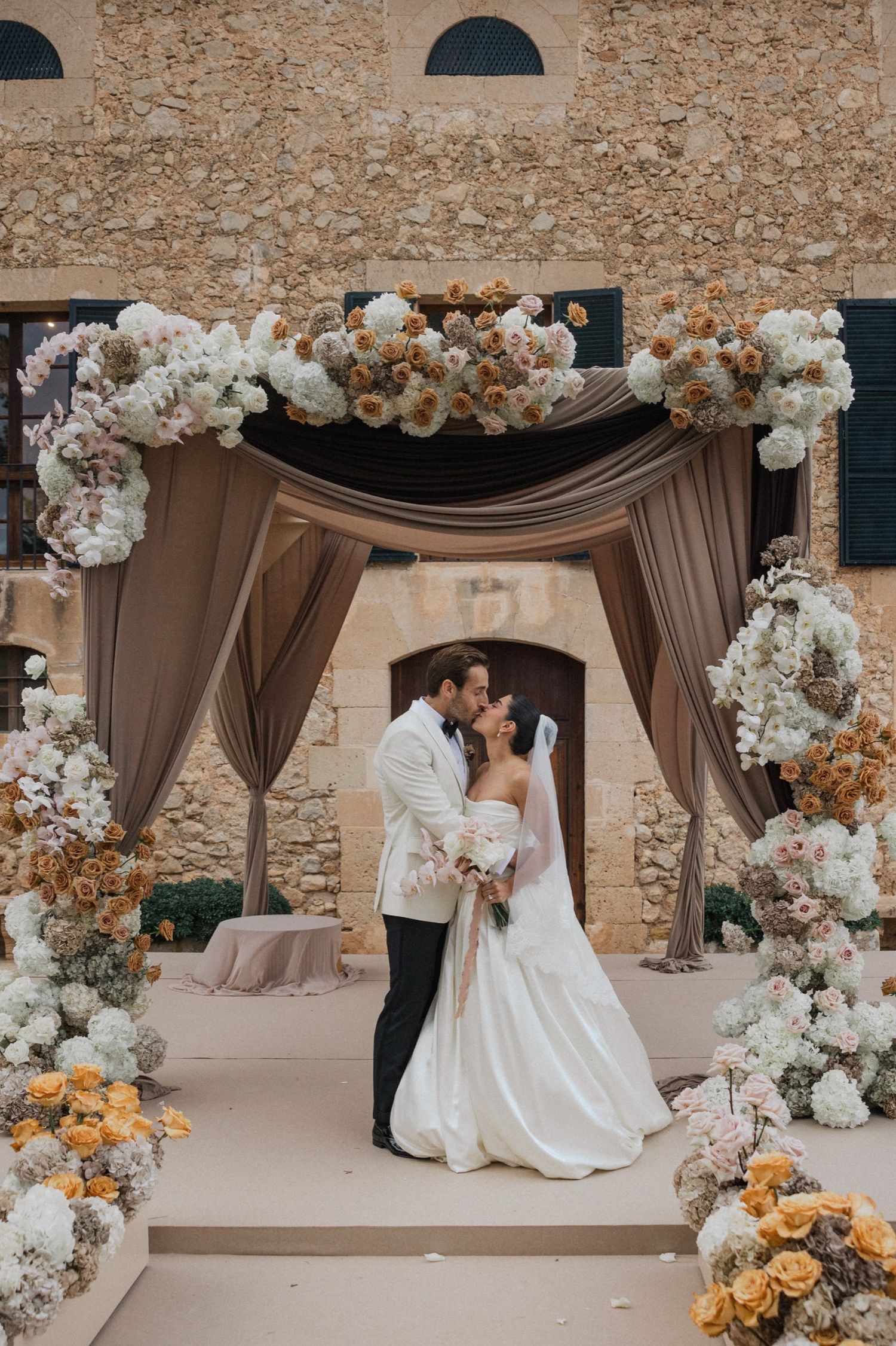 Elegant outdoor wedding ceremony setup with brown drapes and white floral arch decorations against a stone wall.