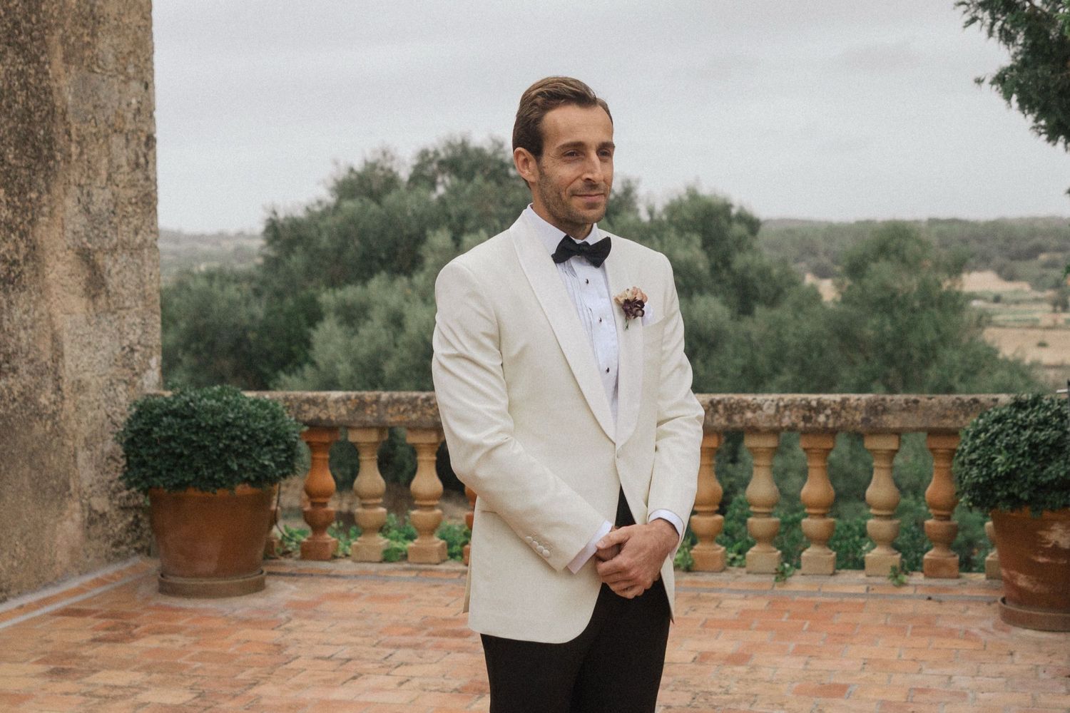 A person in a white tuxedo jacket stands on a rustic terrace with stone columns and Mediterranean landscaping.