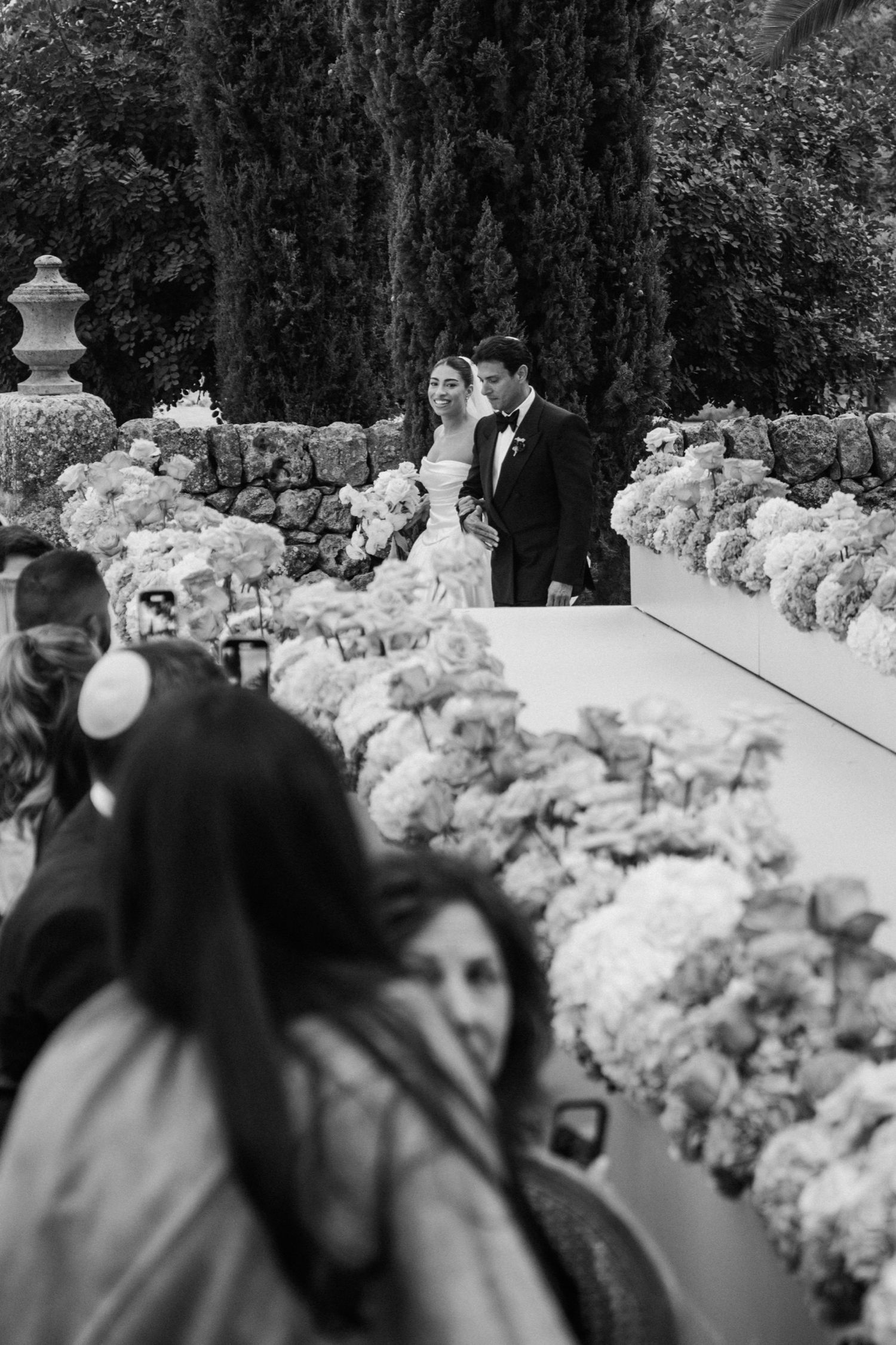 Black and white photo of a wedding ceremony with guests watching and floral arrangements lining the pathway.