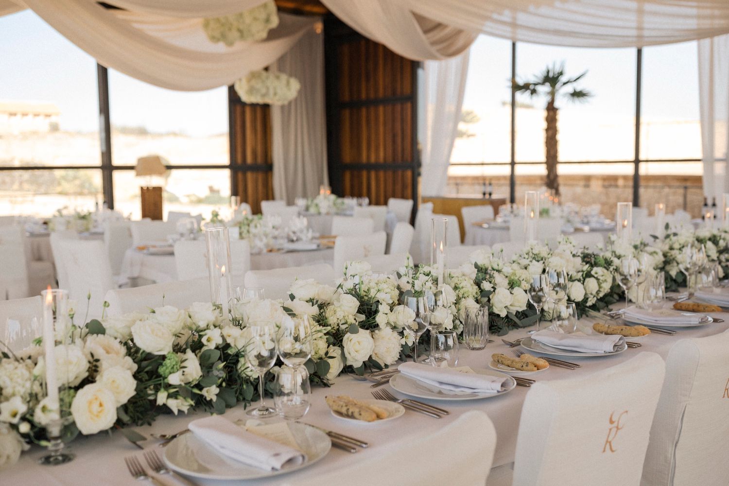 Elegant wedding reception table with white floral centerpieces, candles, and draped ceiling in a sunlit venue.
