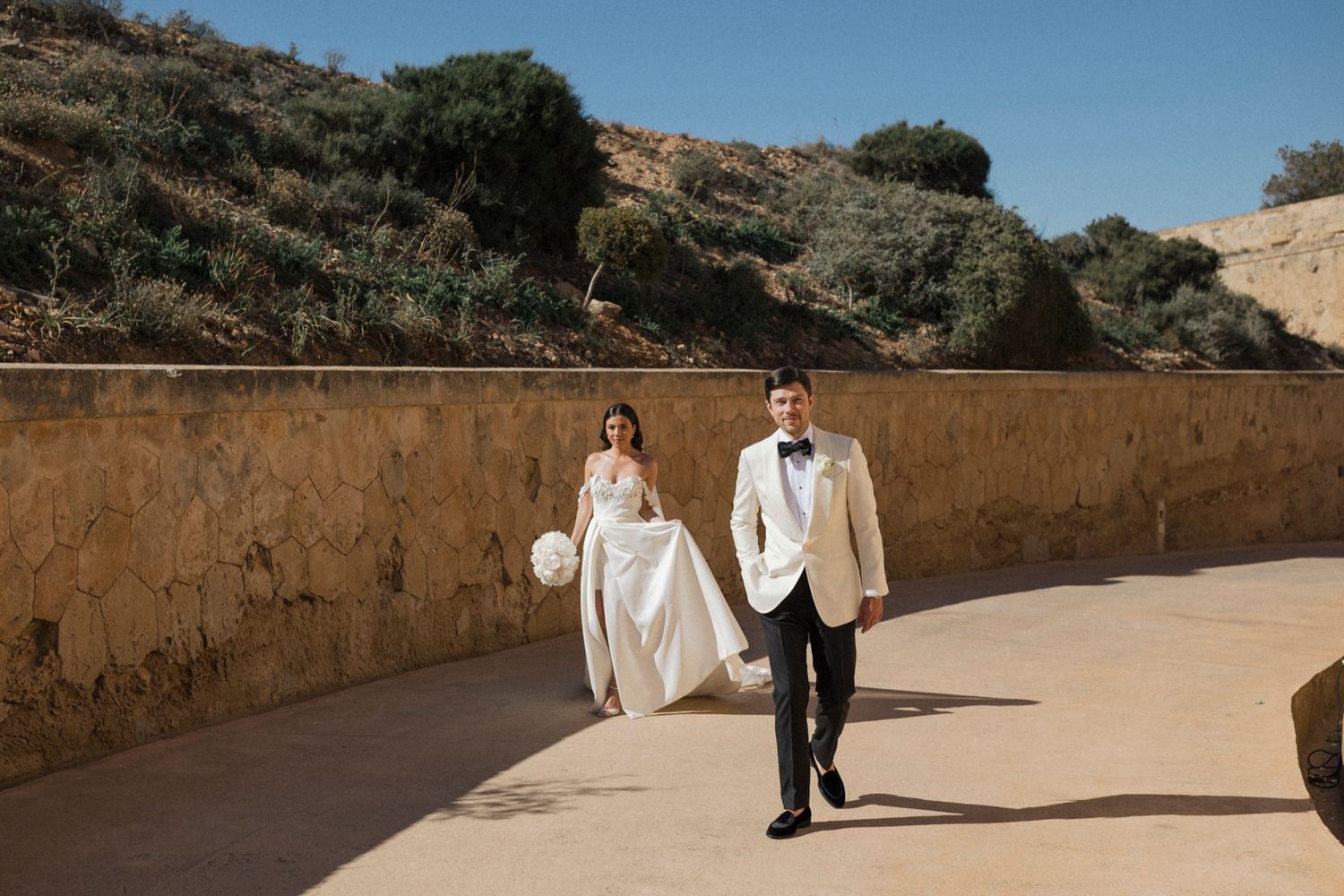 Bride and groom walk hand-in-hand along a sunlit stone pathway with scenic hills in the background.