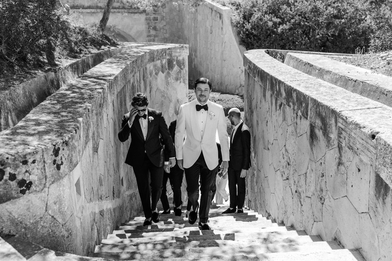 Groom in white tuxedo walks with groomsmen through stone archway steps in elegant black and white wedding photo.