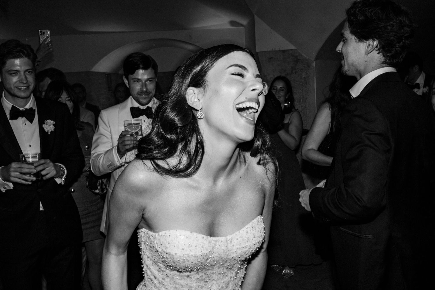 Bride laughing joyfully on wedding dance floor in black and white photo surrounded by guests in formal attire.