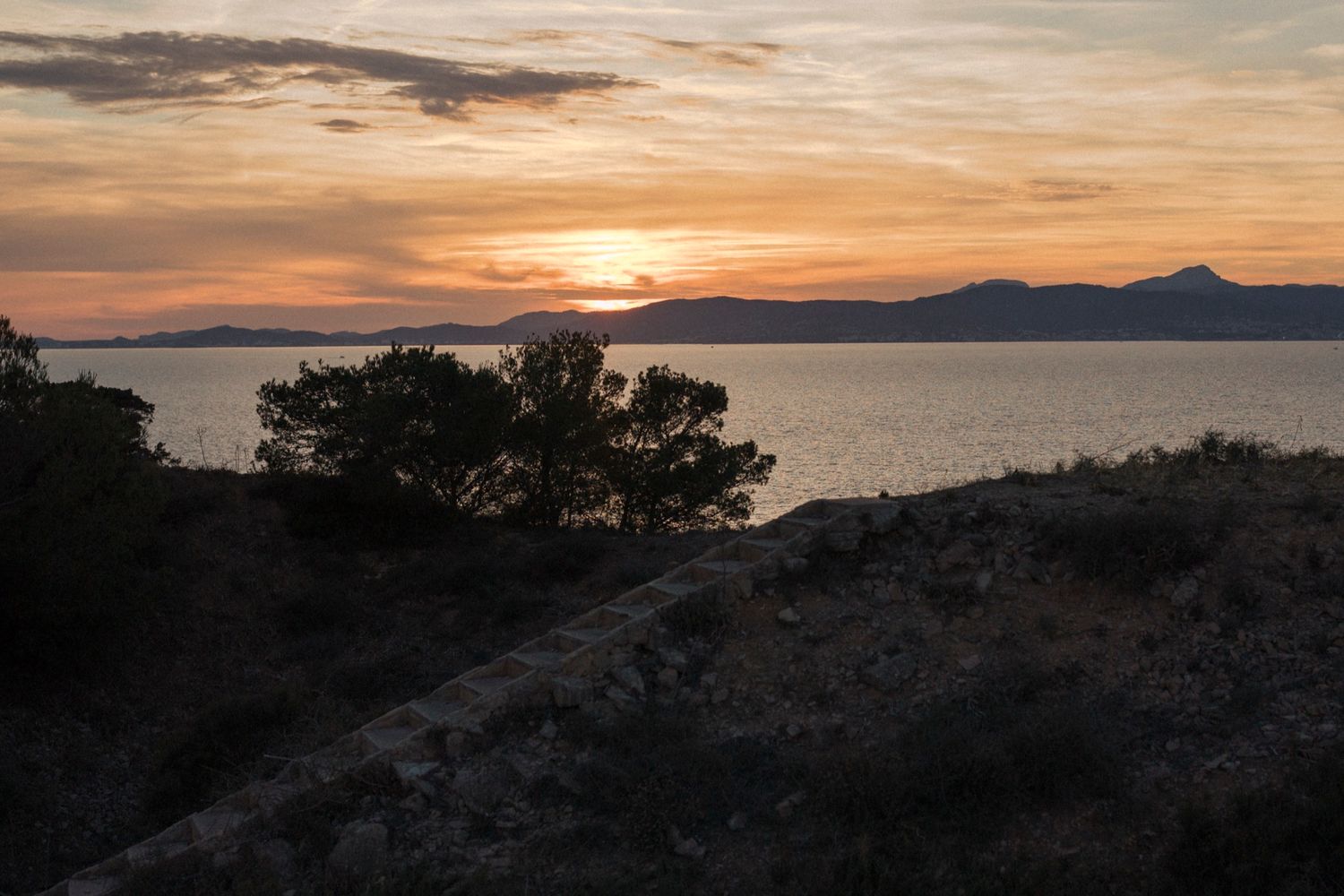 Stunning coastal sunset with silhouetted trees and mountains reflecting warm orange hues over calm Mediterranean waters.