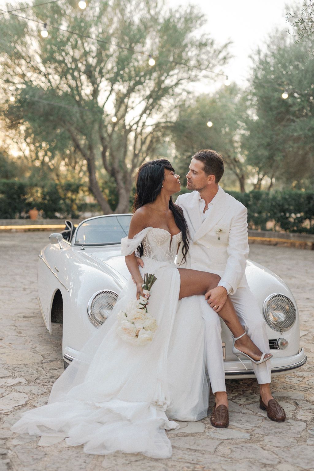 Romantic wedding photoshoot with classic white car in rustic outdoor setting.