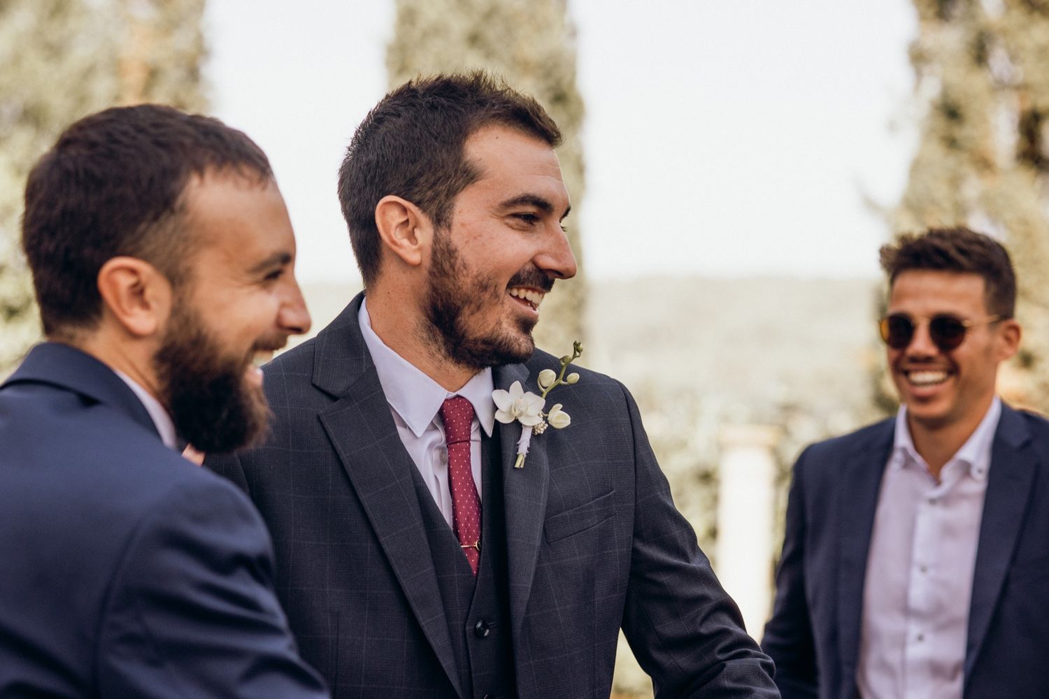 Three men in formal attire share a laugh at an outdoor event.