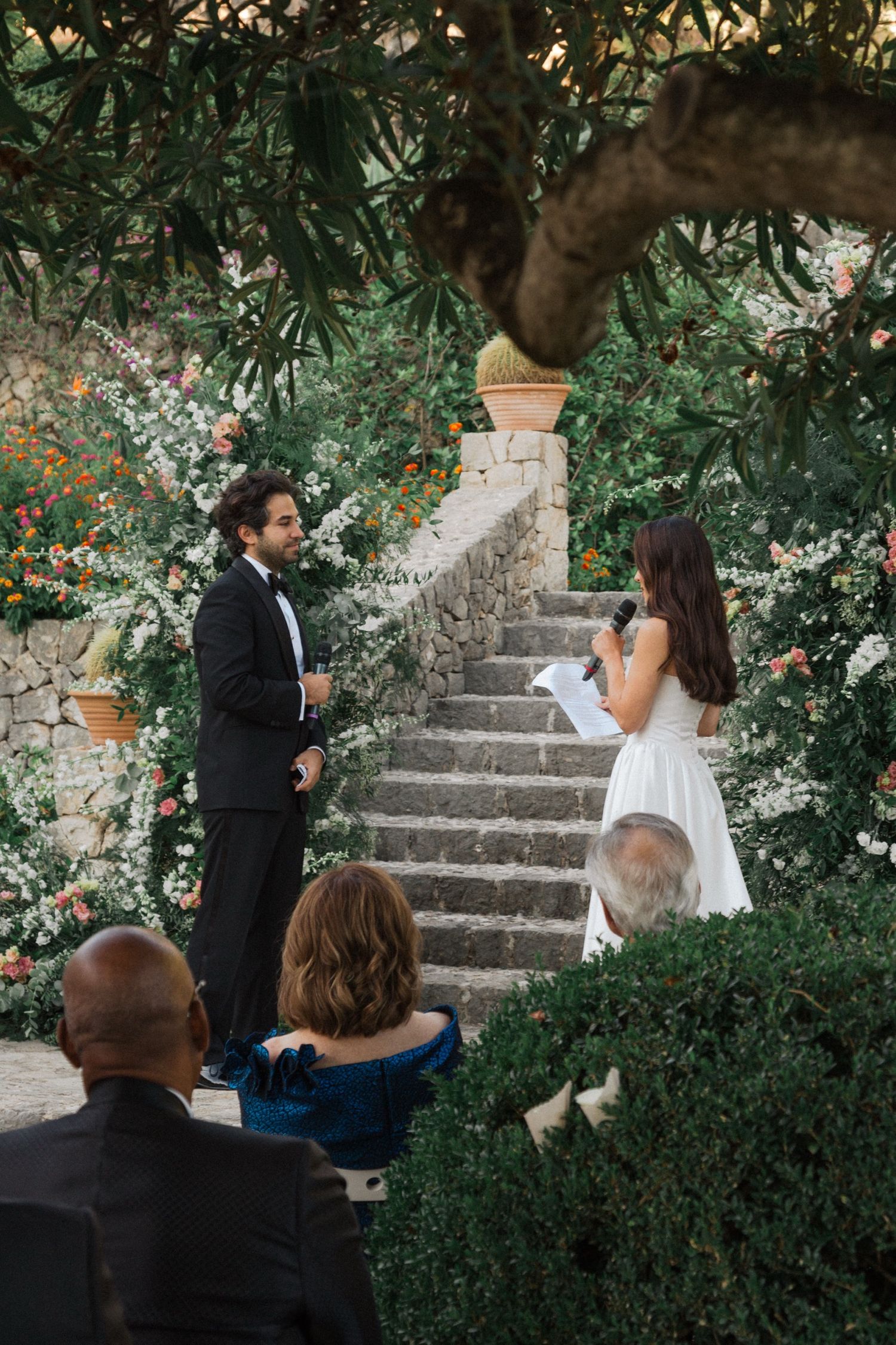 A romantic proposal scene unfolds on garden steps surrounded by blooming flowers and greenery.