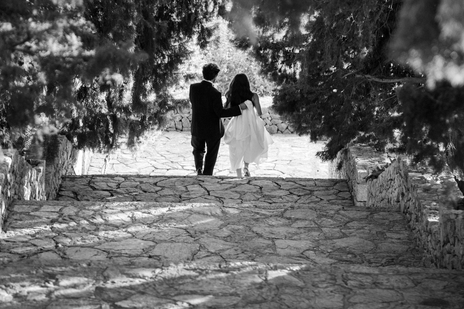 A black and white photograph shows a person walking down a stone path surrounded by trees casting dappled shadows.