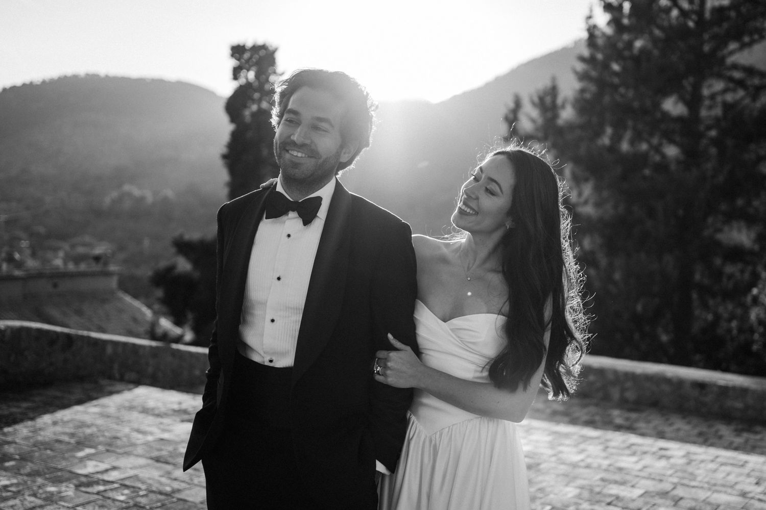 A couple in formal wedding attire shares a romantic moment on a mountaintop at sunset.