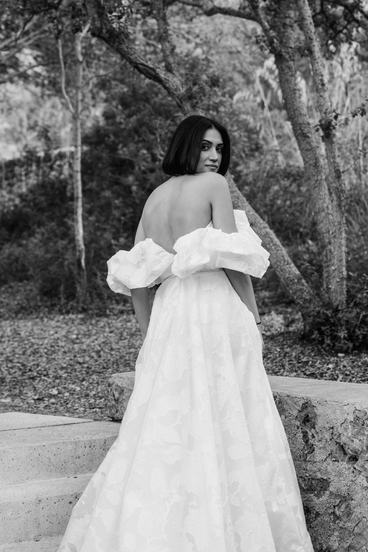 A person in a flowing white off-shoulder dress poses in a natural outdoor setting with trees in the background.
