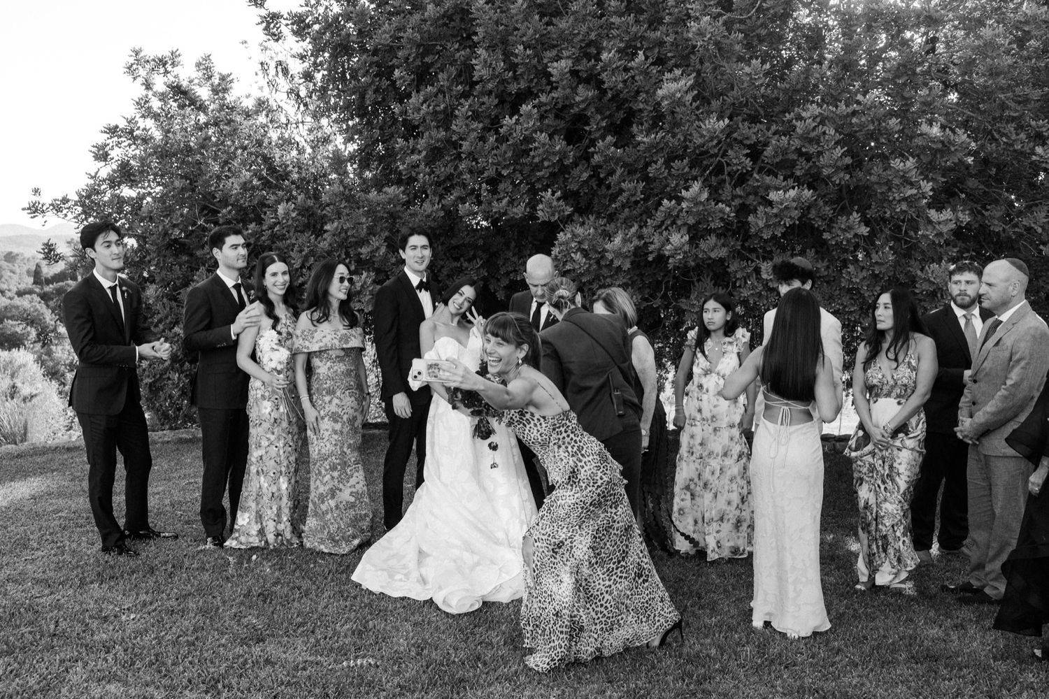 Wedding party poses outdoors under trees in formal attire during an elegant garden ceremony.