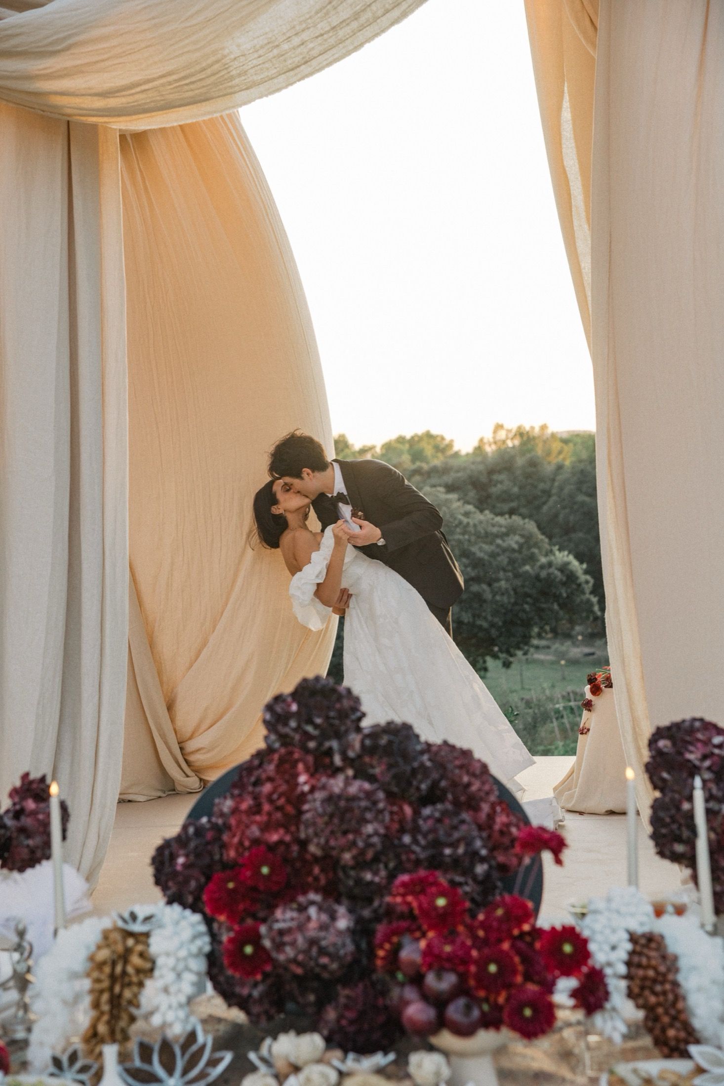 A romantic wedding couple shares a kiss under flowing cream drapery with deep burgundy floral arrangements in the foreground.