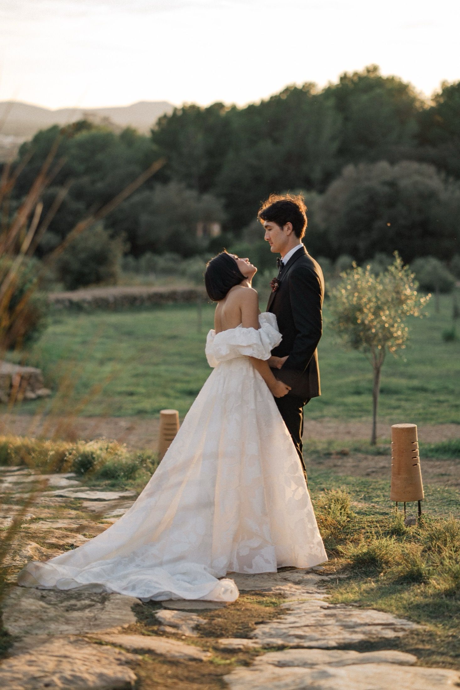 A couple in wedding attire embraces during golden hour in a scenic outdoor setting with flowing white dress.