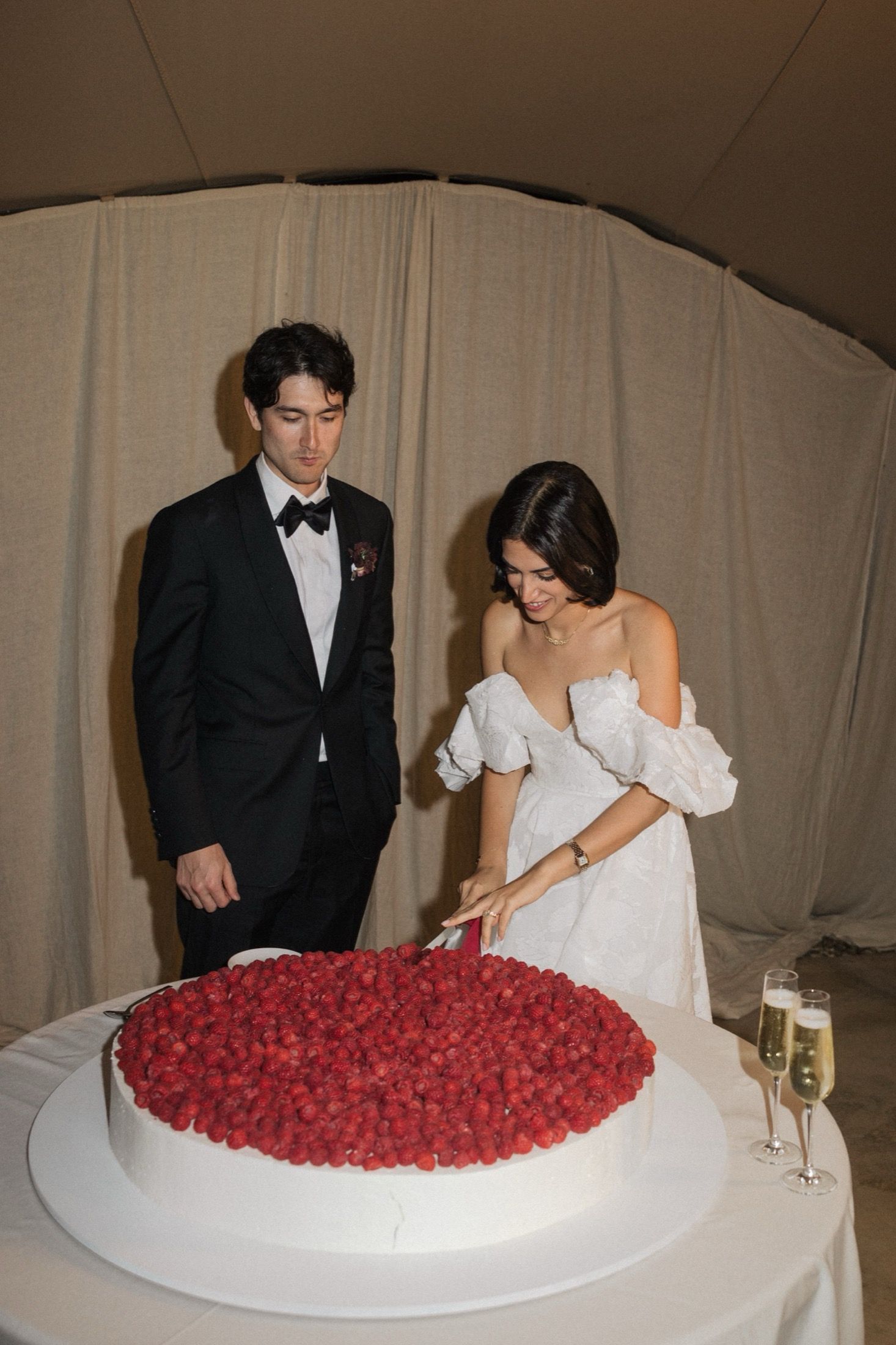 A couple in formal wedding attire cuts into a large white cake decorated with fresh red raspberries.
