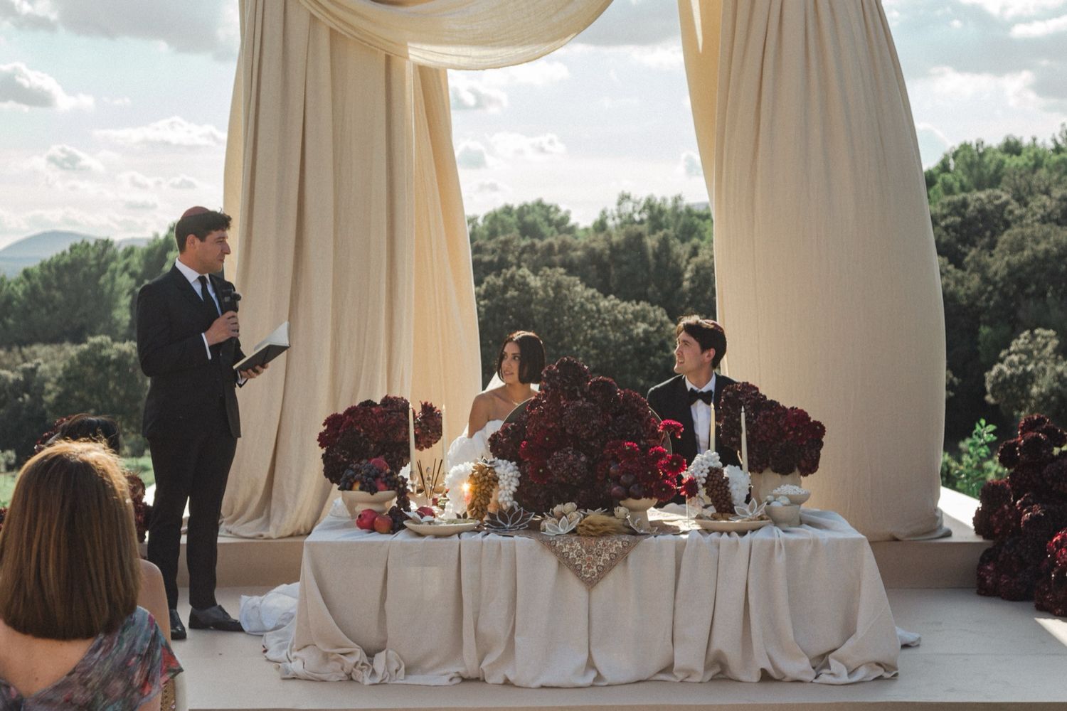 An outdoor wedding reception setup with white draping, red floral centerpieces and guests seated at an elegant table.