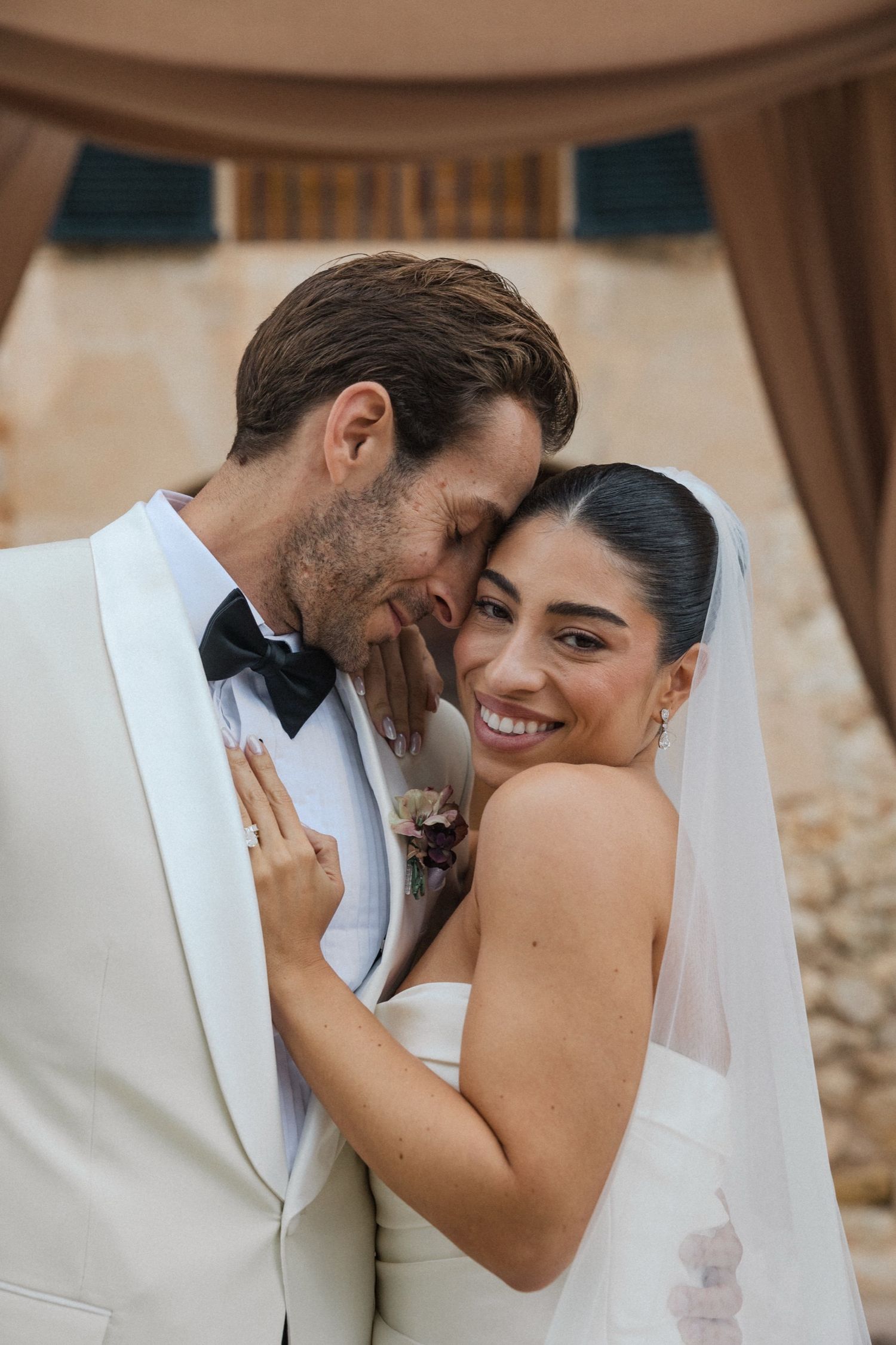A romantic wedding moment captured with a bride in white veil and groom in white tuxedo embracing and smiling.