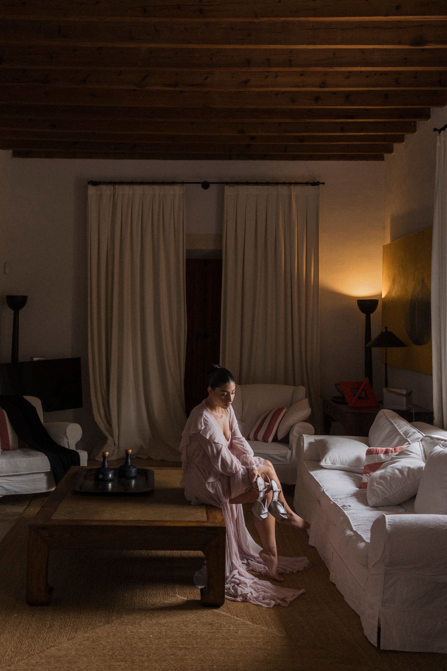 Soft lighting illuminates a cozy living room with white furniture and exposed wooden beams.