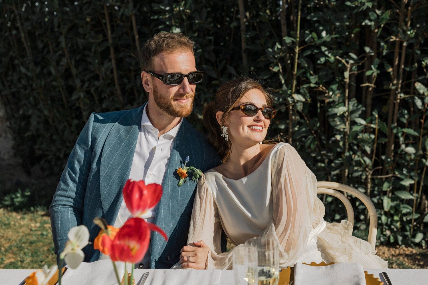 A couple enjoys cocktails and conversation in an outdoor garden setting with natural sunlight filtering through trees.
