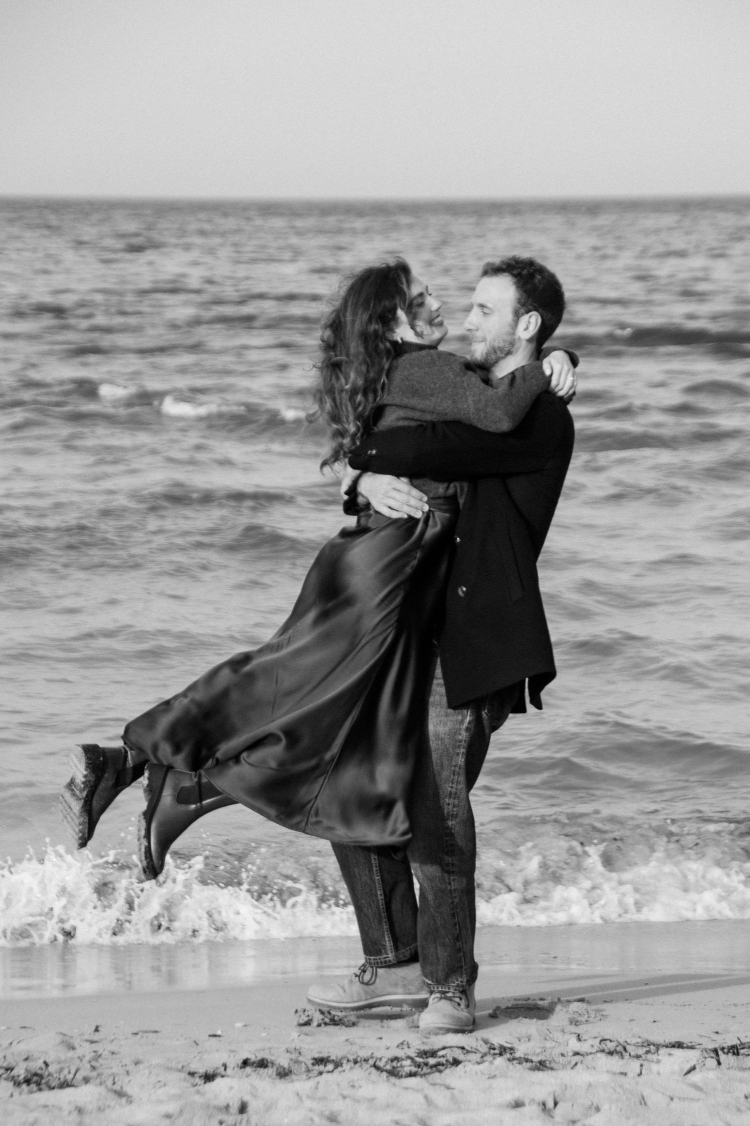 A romantic black and white photo of a couple embracing on a beach with waves crashing behind them.