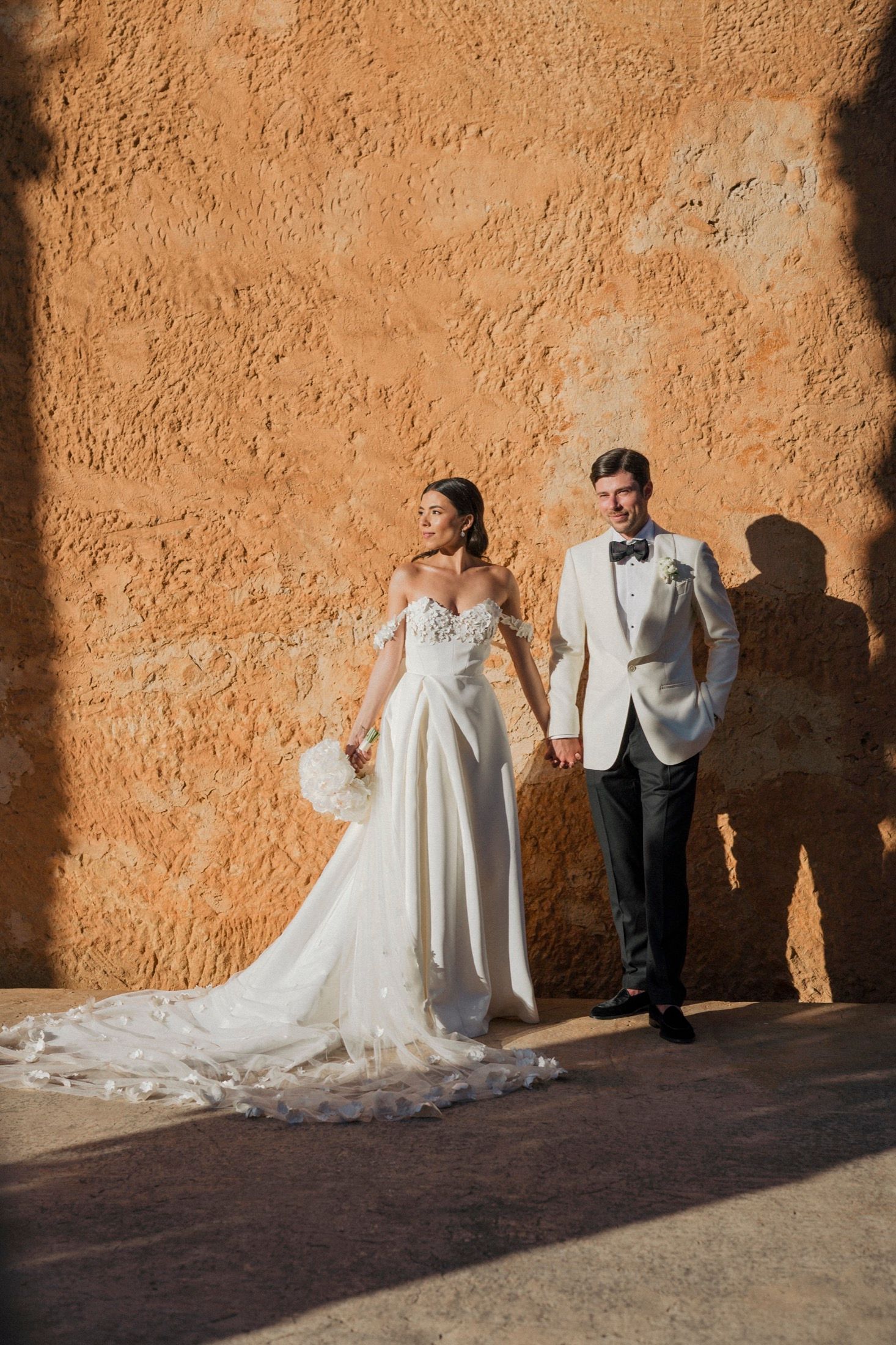 A bride in a white gown and groom in a white tuxedo hold hands against a warm terracotta wall in sunlight.