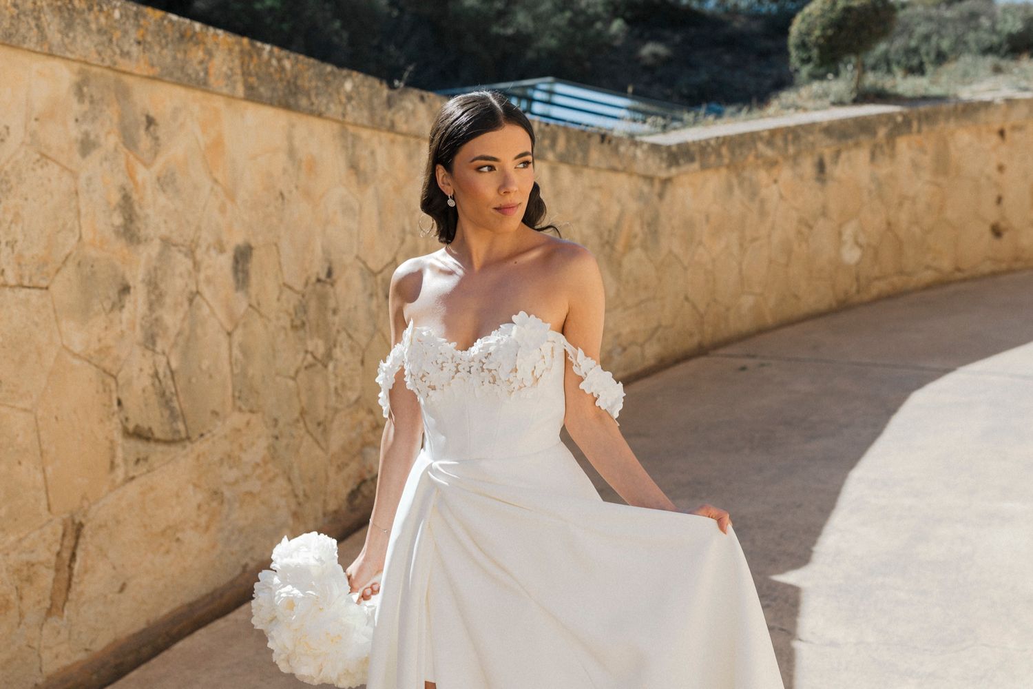 Bride in elegant off-shoulder white wedding gown with floral details holds bouquet outdoors near stone wall.