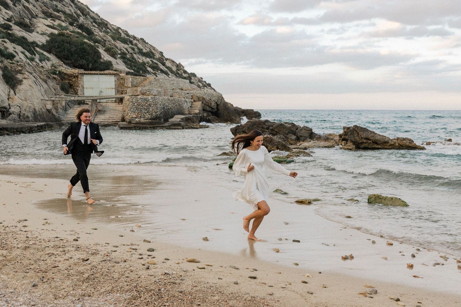 Couple walking along a dramatic rocky coastline with waves crashing on a cloudy day.