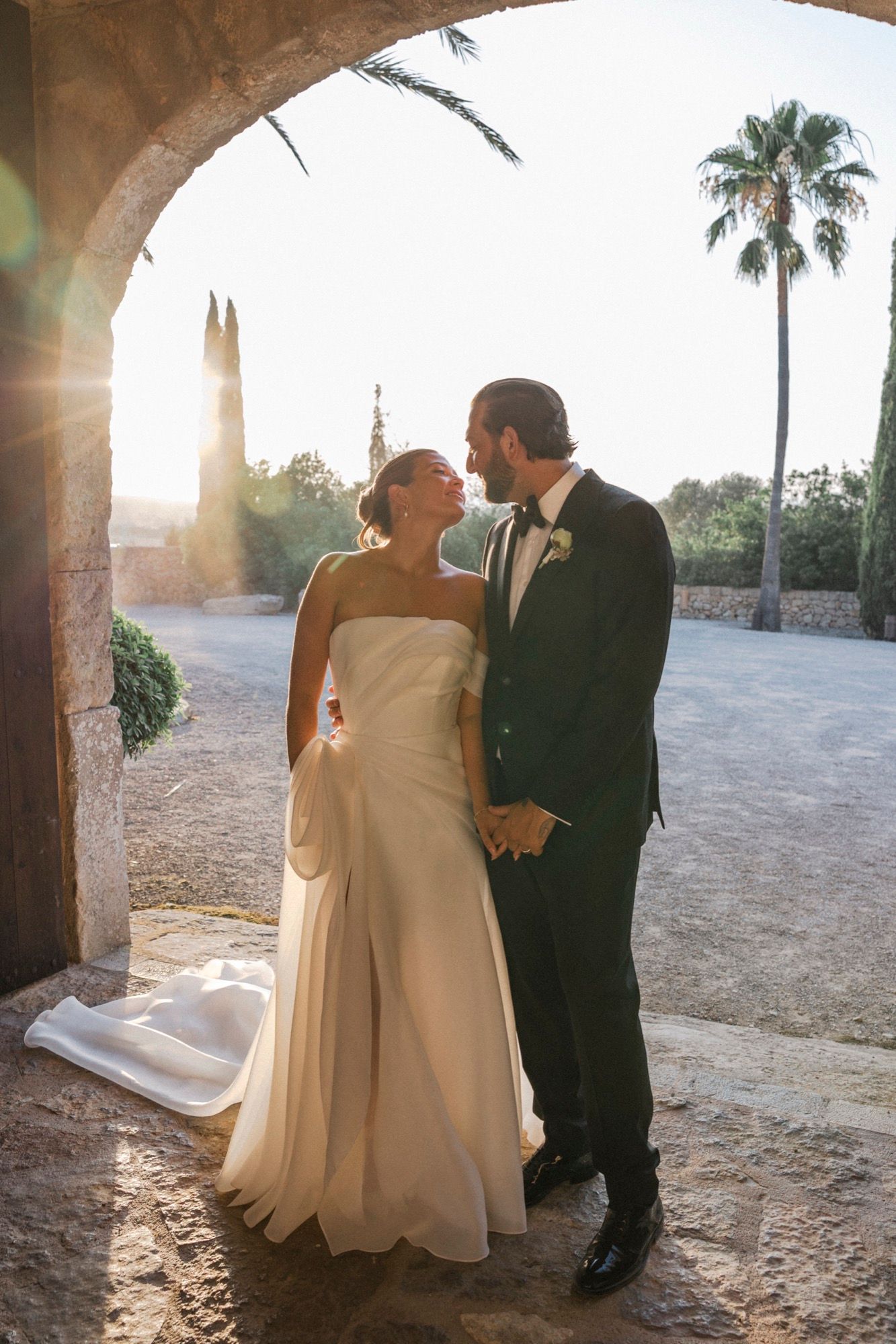 A newlywed couple shares an intimate moment under a stone archway at sunset with a palm tree in view.