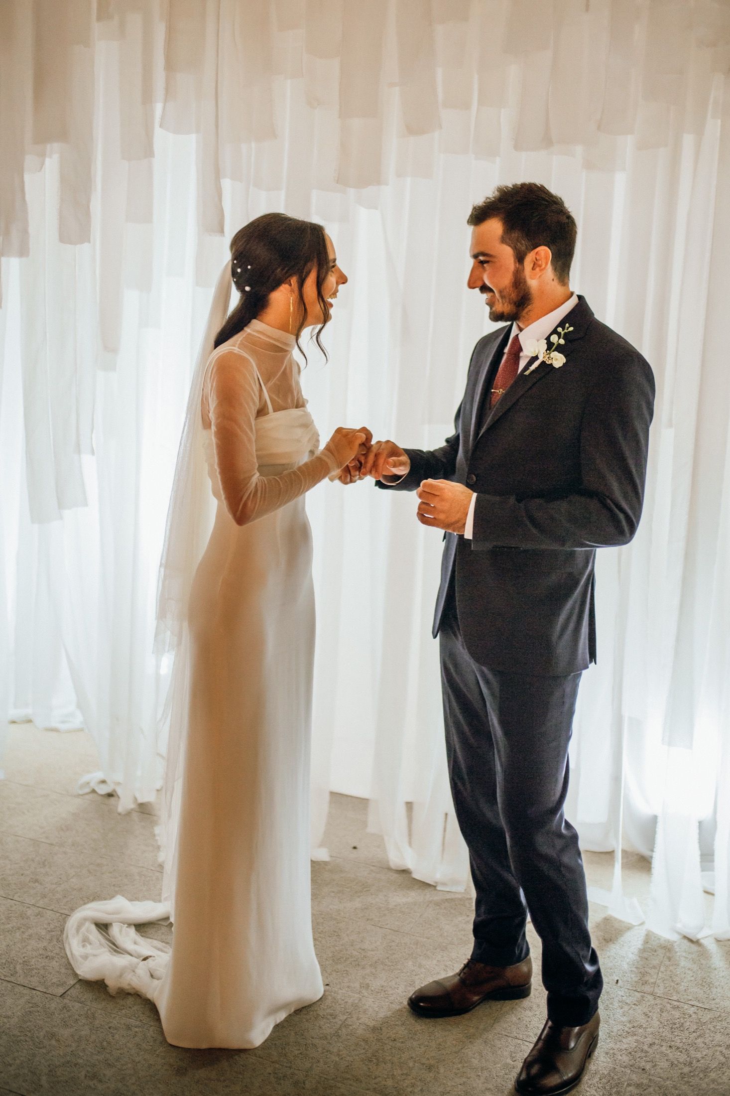 A bride and groom exchange rings during an intimate wedding ceremony against white curtained backdrop.