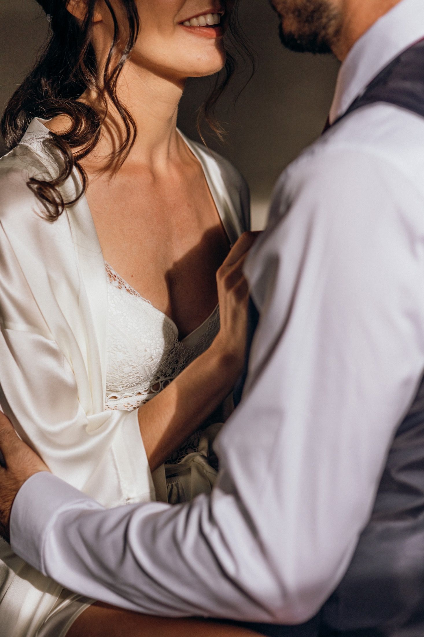 Close-up detail of a bride in a white satin robe and groom in a dark suit embracing during an intimate wedding moment.